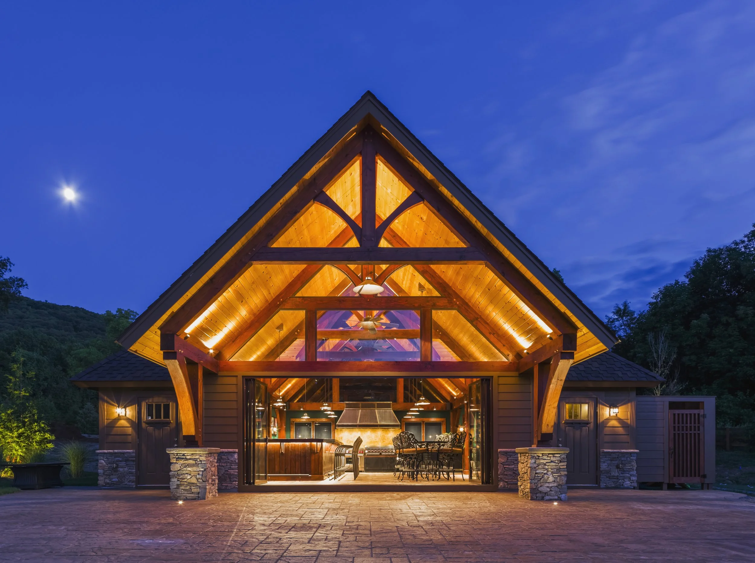 Striking timber-frame lodge bar exterior at night, warmly illuminated with golden interior lighting that glows through the vaulted ceiling and glass panels, contrasting against the deep blue evening sky and moonlight.