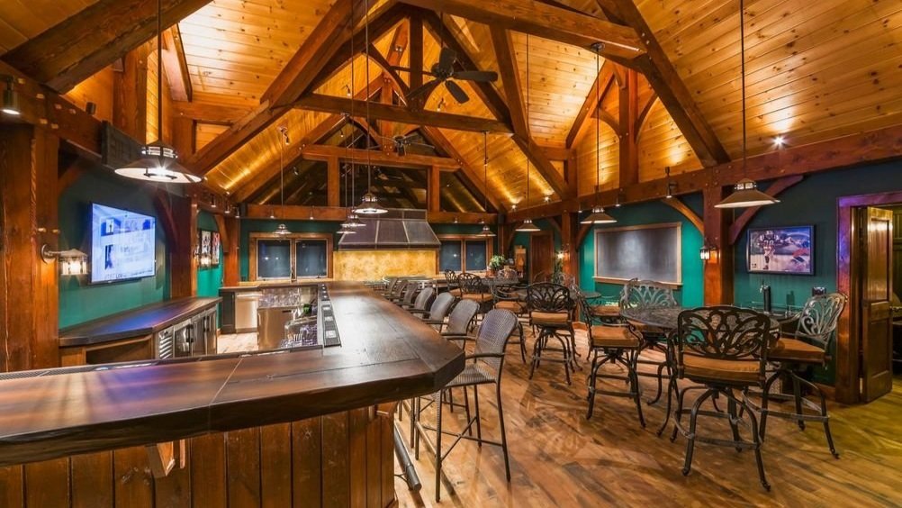 Spacious rustic lodge bar with exposed wooden beams, high vaulted ceilings, and warm ambient lighting from pendant lamps and ceiling fixtures, creating a cozy and inviting atmosphere.
