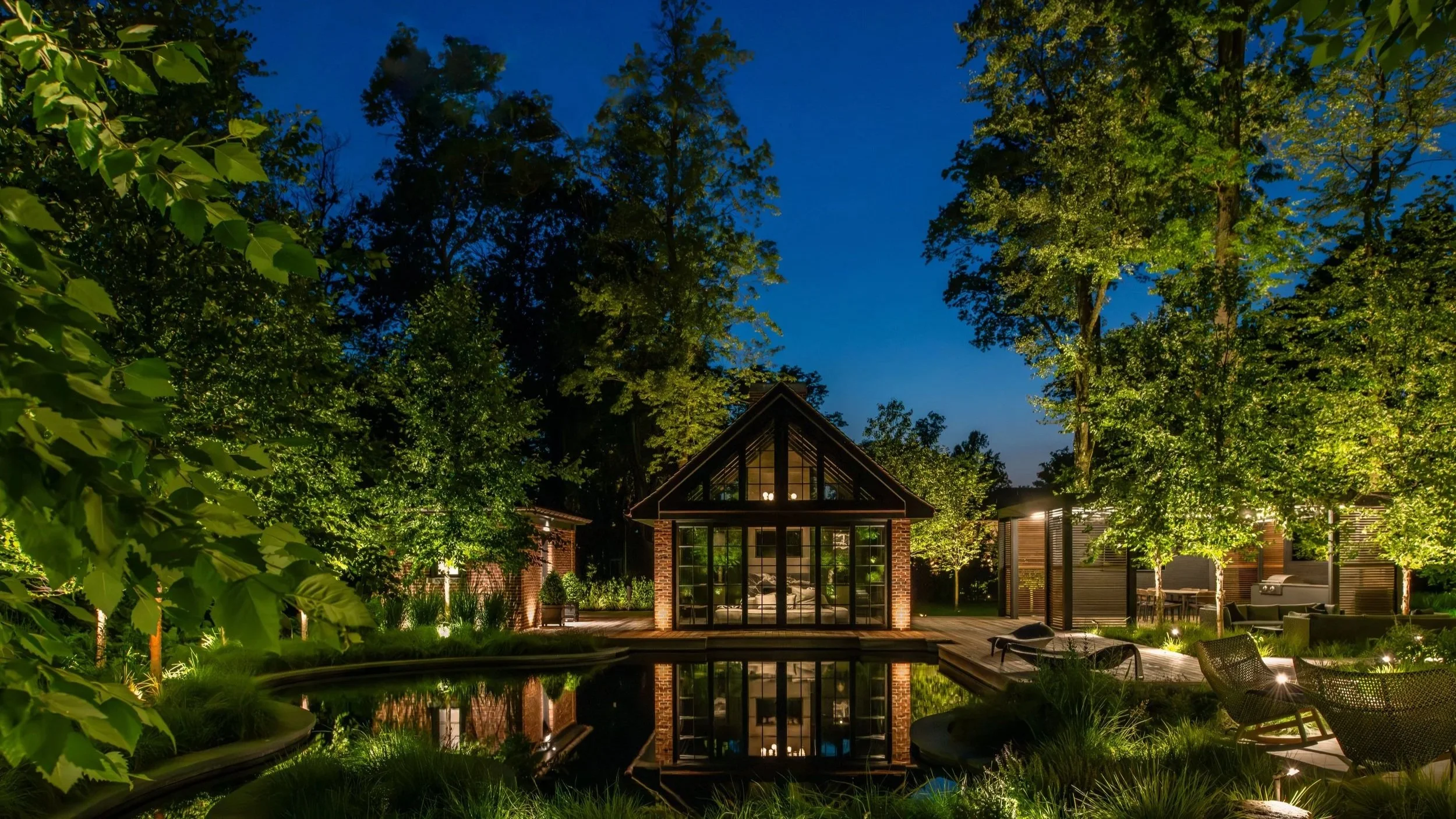 A softly illuminated water feature leading toward a modern home, framed by warm LED tree uplighting and layered planting across a New Jersey estate at night.