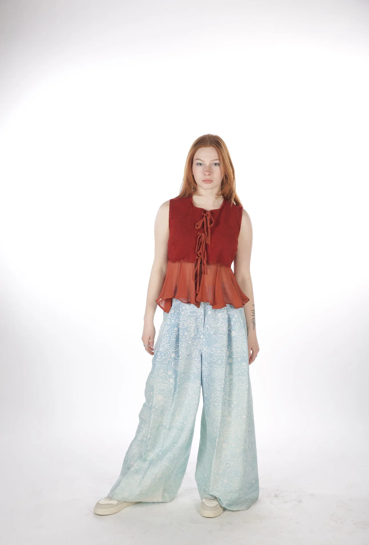 Red-haired woman wearing a sleeveless red and orange top with a bow detail, wide-leg patterned light blue pants, and sneakers, standing against a white background.