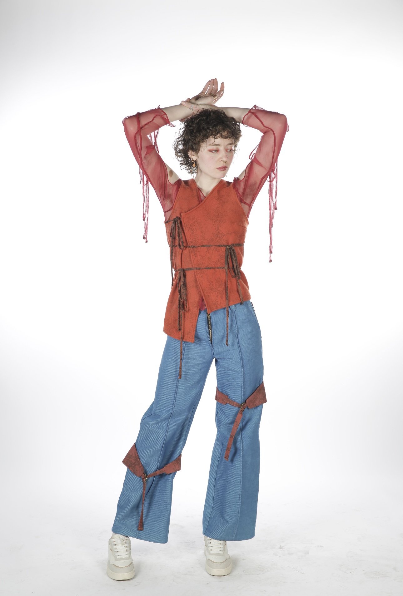 A young woman with short curly hair wearing a red layered top with sheer sleeves and blue wide-leg pants with red straps, standing against a white background with her arms raised above her head.