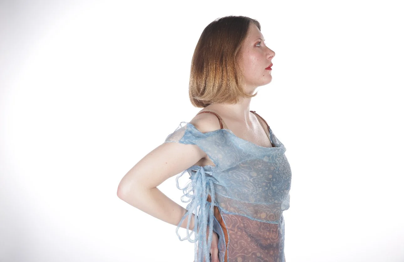 Profile of a woman with shoulder-length brown hair wearing a sheer blue dress with lace details, standing with hands behind her back against a white background.
