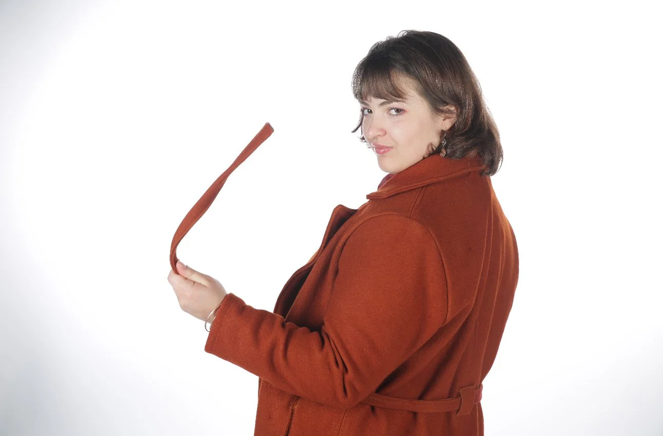 Woman with shoulder-length brown hair wearing a rust-colored coat, holding a brown leather belt, standing against a white background.