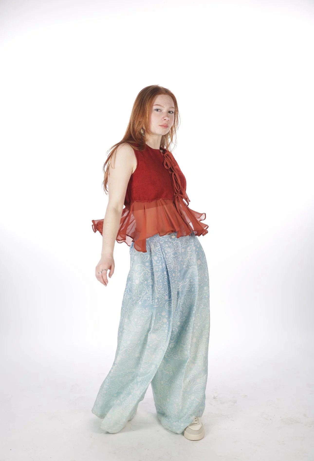 A young woman with red hair wearing a red sleeveless top and wide-leg light blue patterned pants standing against a white background.