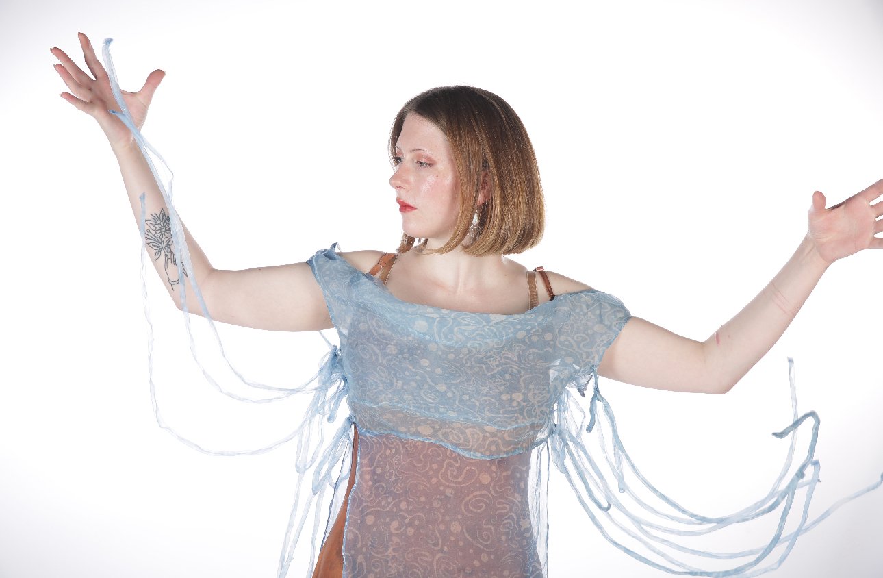 A woman with short brown hair wearing a sheer, light blue dress with flowing ribbon-like sleeves, standing against a white background, with her arms extended outward.