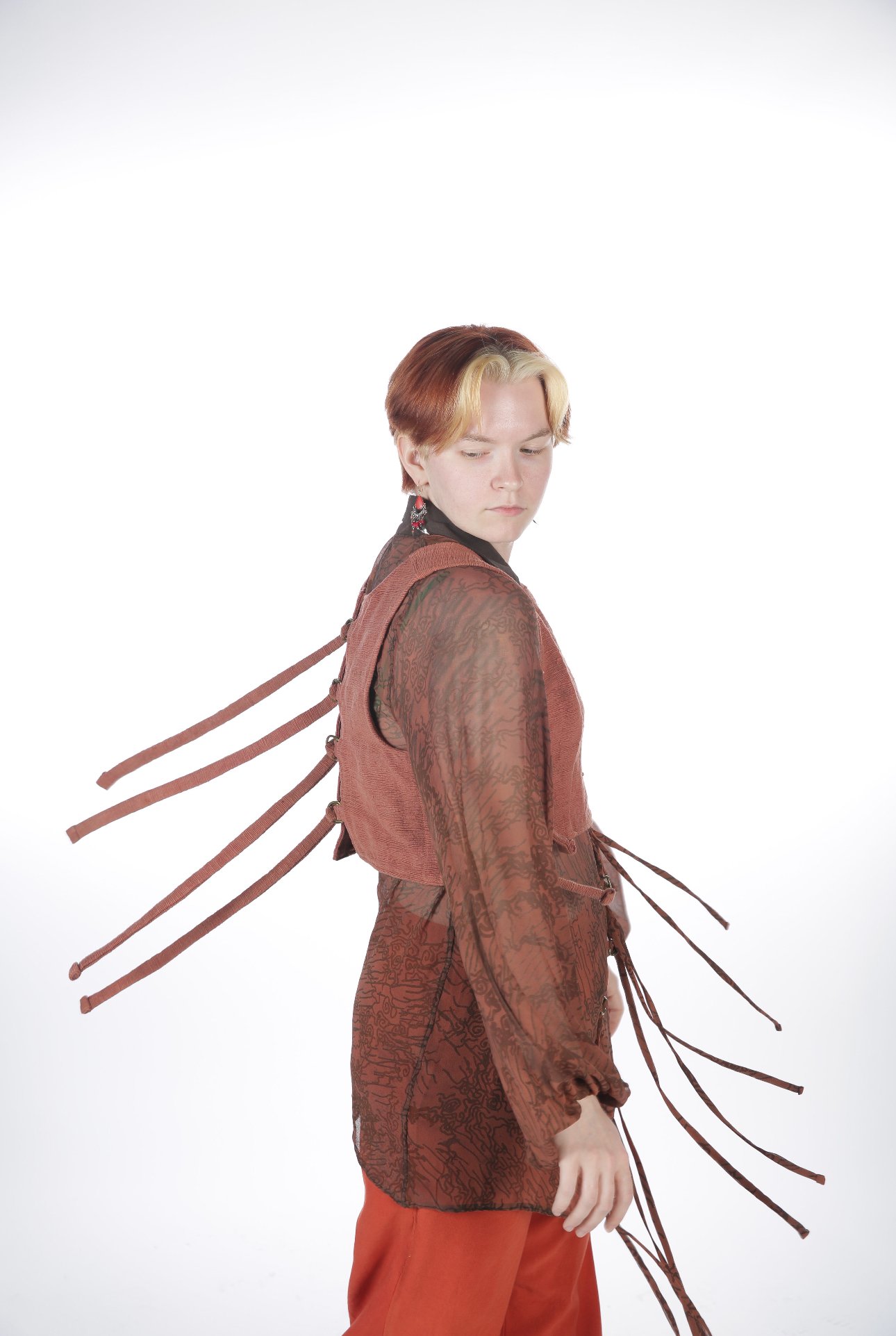 A young person with short, two-toned hair wearing a sheer, long-sleeved top, a vest with multiple straps, and matching pants, posing against a plain white background.