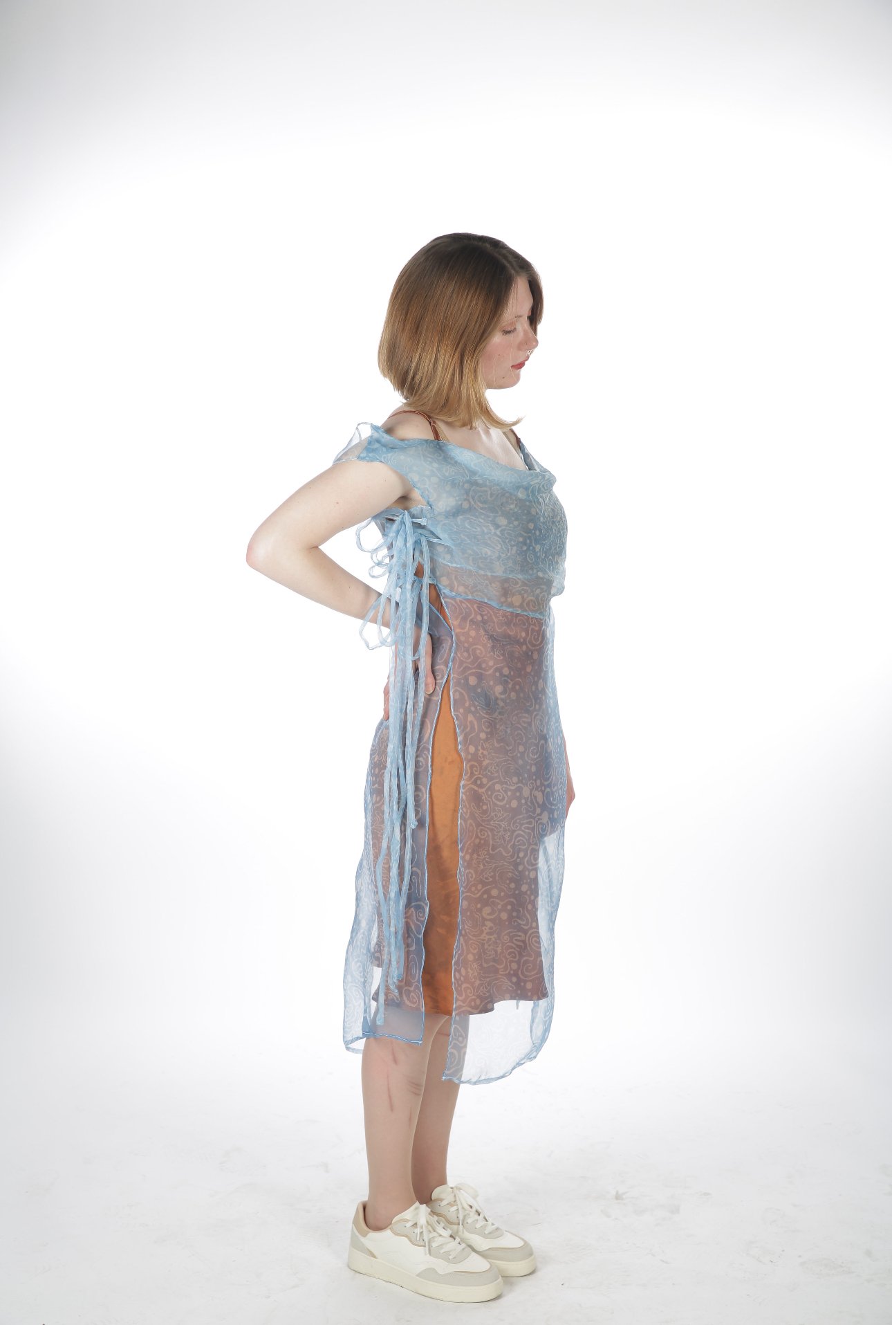 A woman wearing a sheer blue dress with side slits, standing sideways with her hands behind her back on a white background.