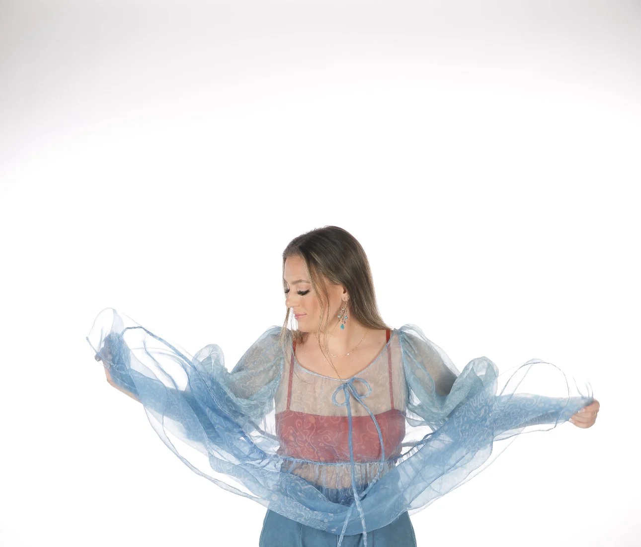 A woman with long brown hair wearing a sheer blue and red top, holding the blue fabric of her sheer sleeves out to the sides against a white background.