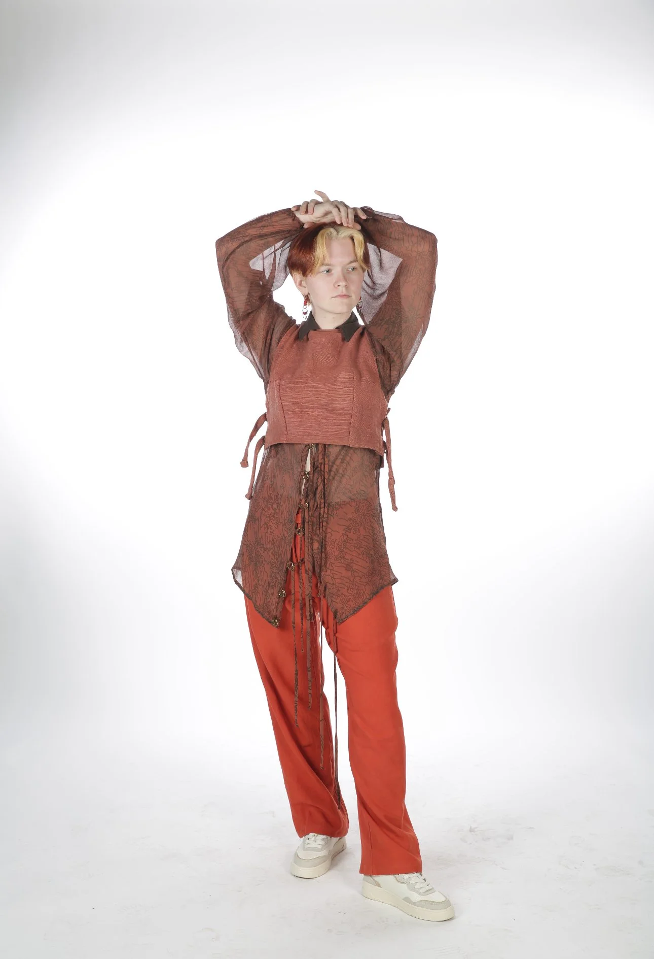 Person with short hair wearing a layered rust-colored outfit and white sneakers, standing with arms raised and crossed behind their head, against a plain white background.