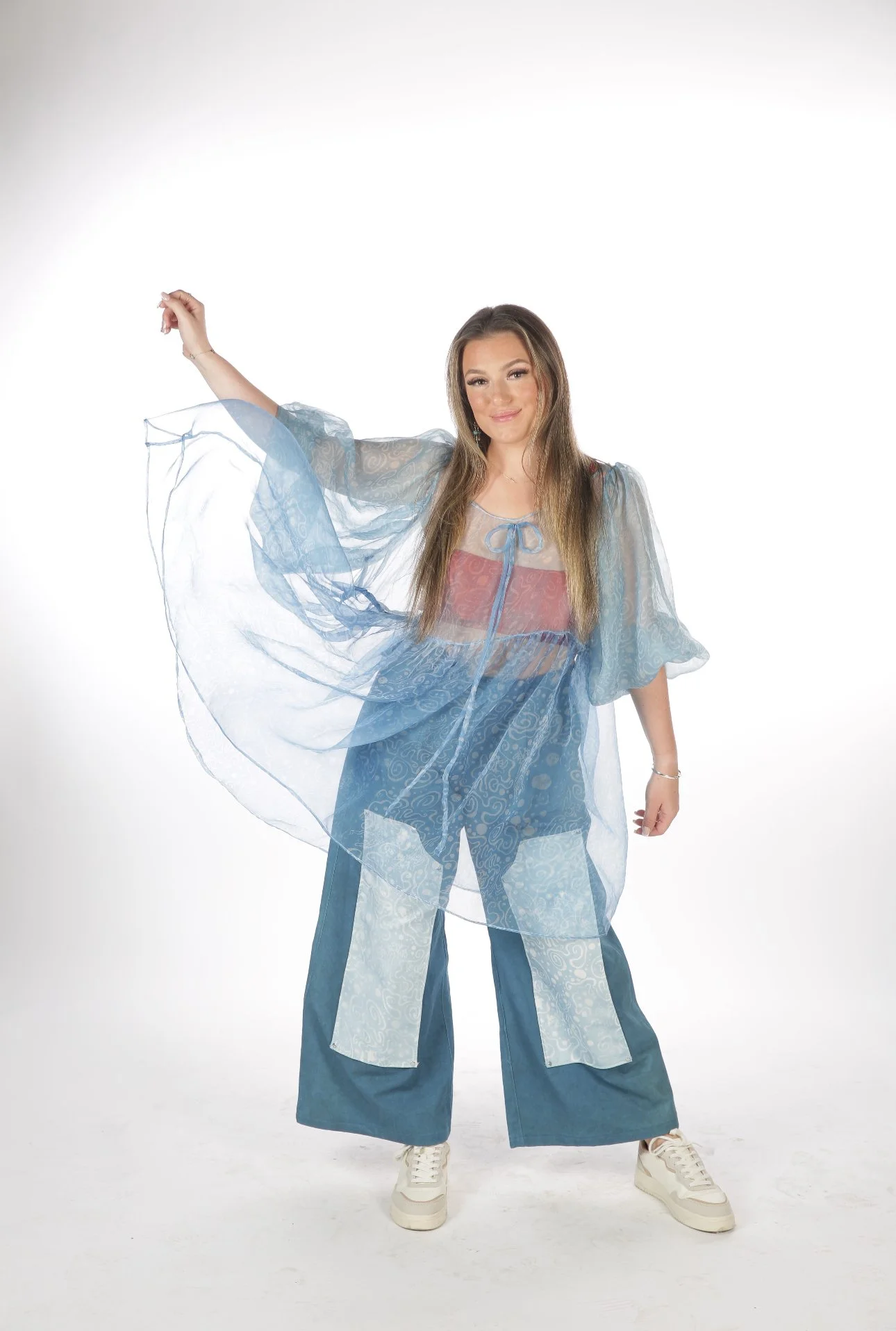 A young woman wearing a blue sheer layered dress with wide-leg pants, standing on a white background, smiling and posing with one arm raised.