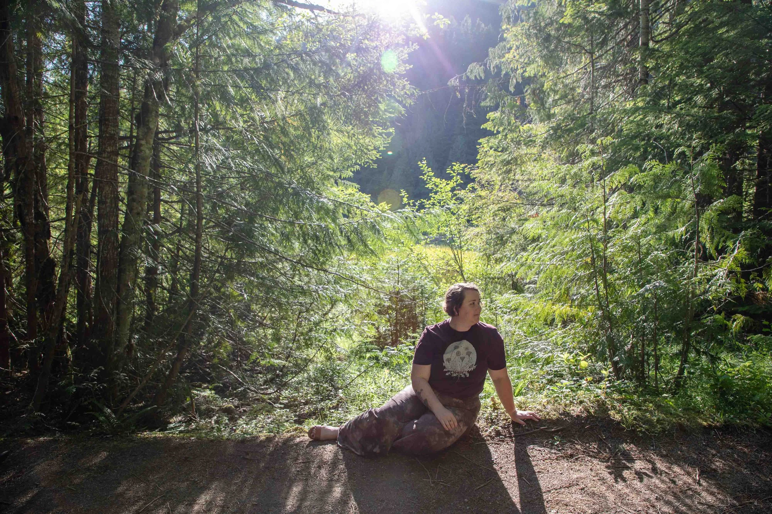 A woman sitting on a dirt path in a sunlit forest surrounded by tall green trees.