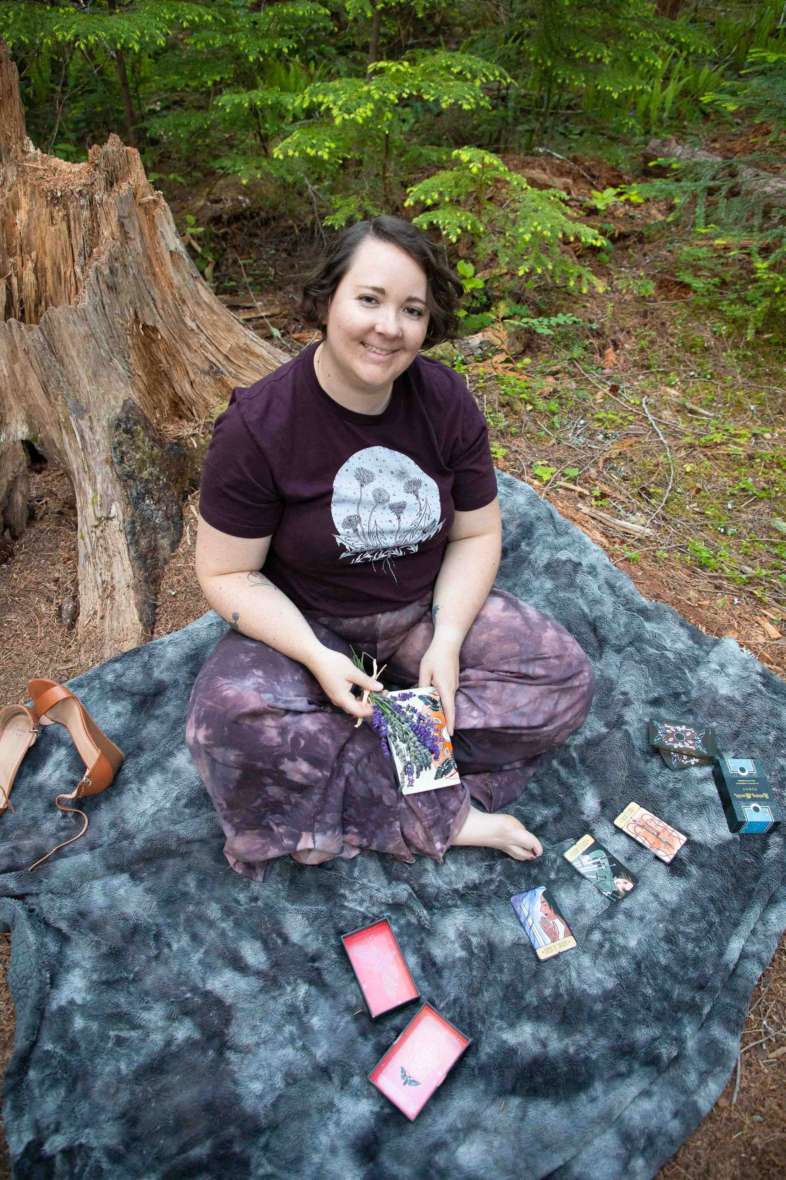 A woman sits cross-legged on a black furry blanket outdoors in a wooded area, holding a booklet and surrounded by tarot cards and card boxes, smiling at the camera.