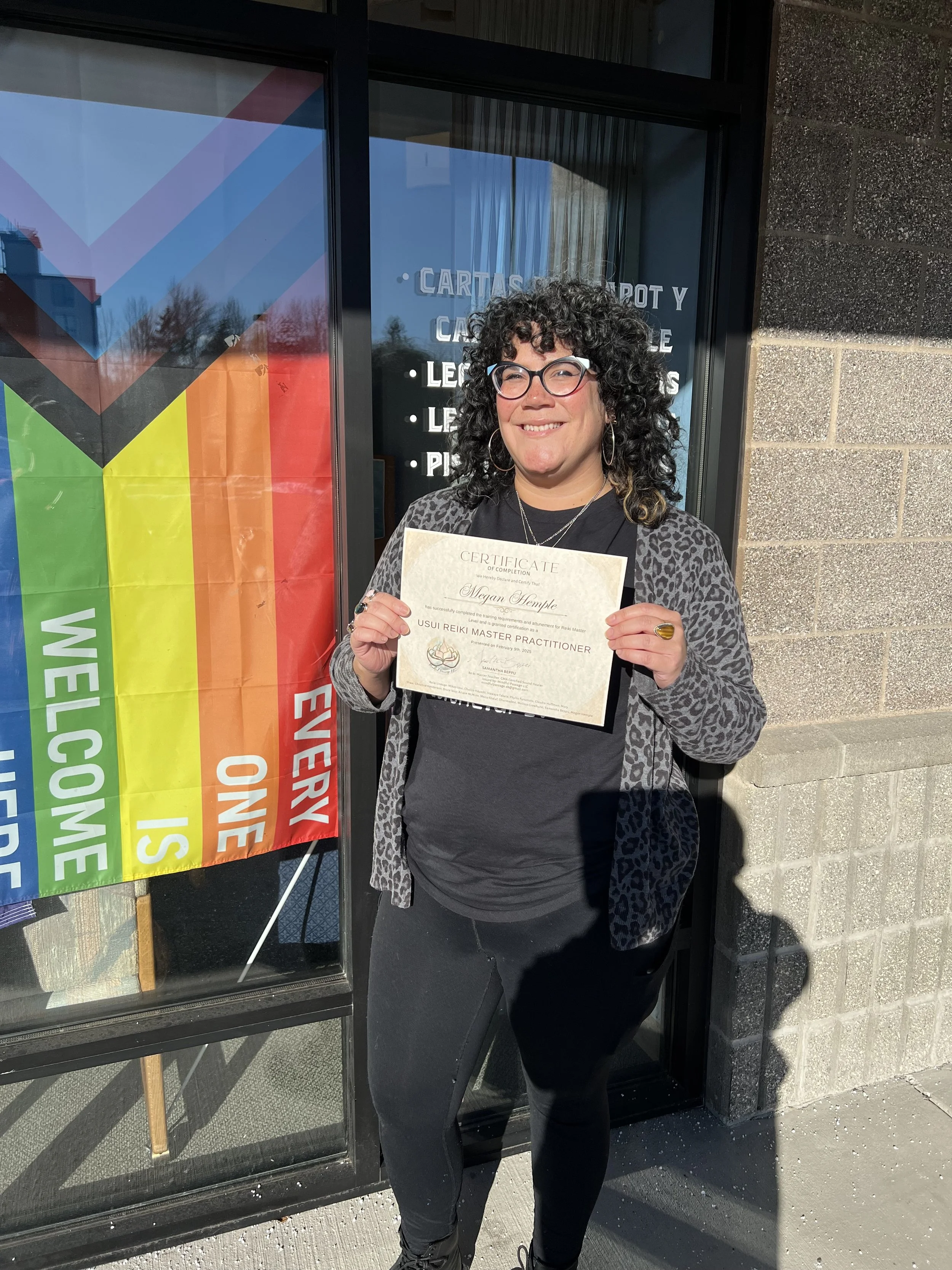 Woman with curly dark hair and glasses, smiling, holding a certificate in front of a store window. She is dressed in a black shirt, dark pants, and a patterned cardigan. The store window displays a rainbow flag and a sign with colorful text.