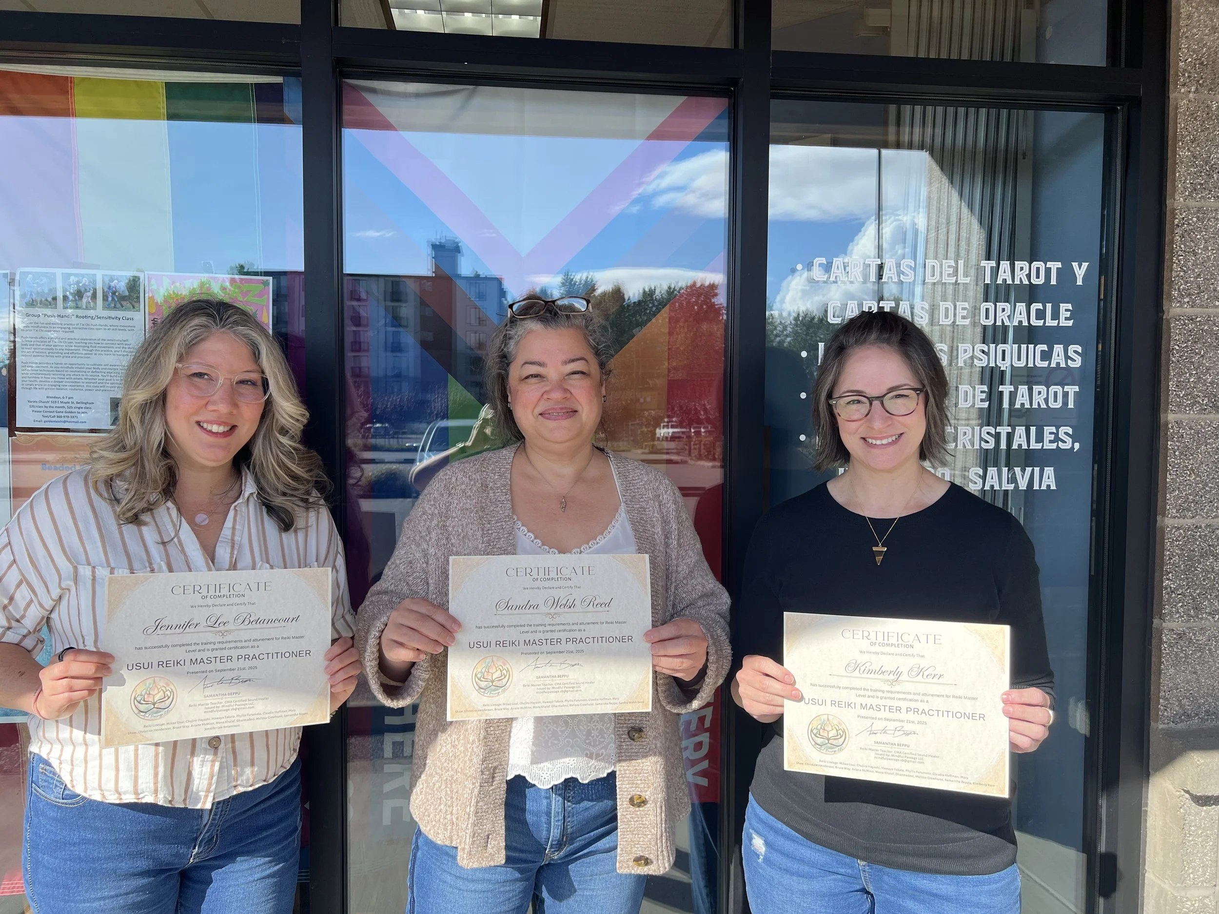 Three women standing outside a storefront holding certificates, smiling at the camera. The store has a glass door with signs in Spanish and visible reflections of buildings and trees.