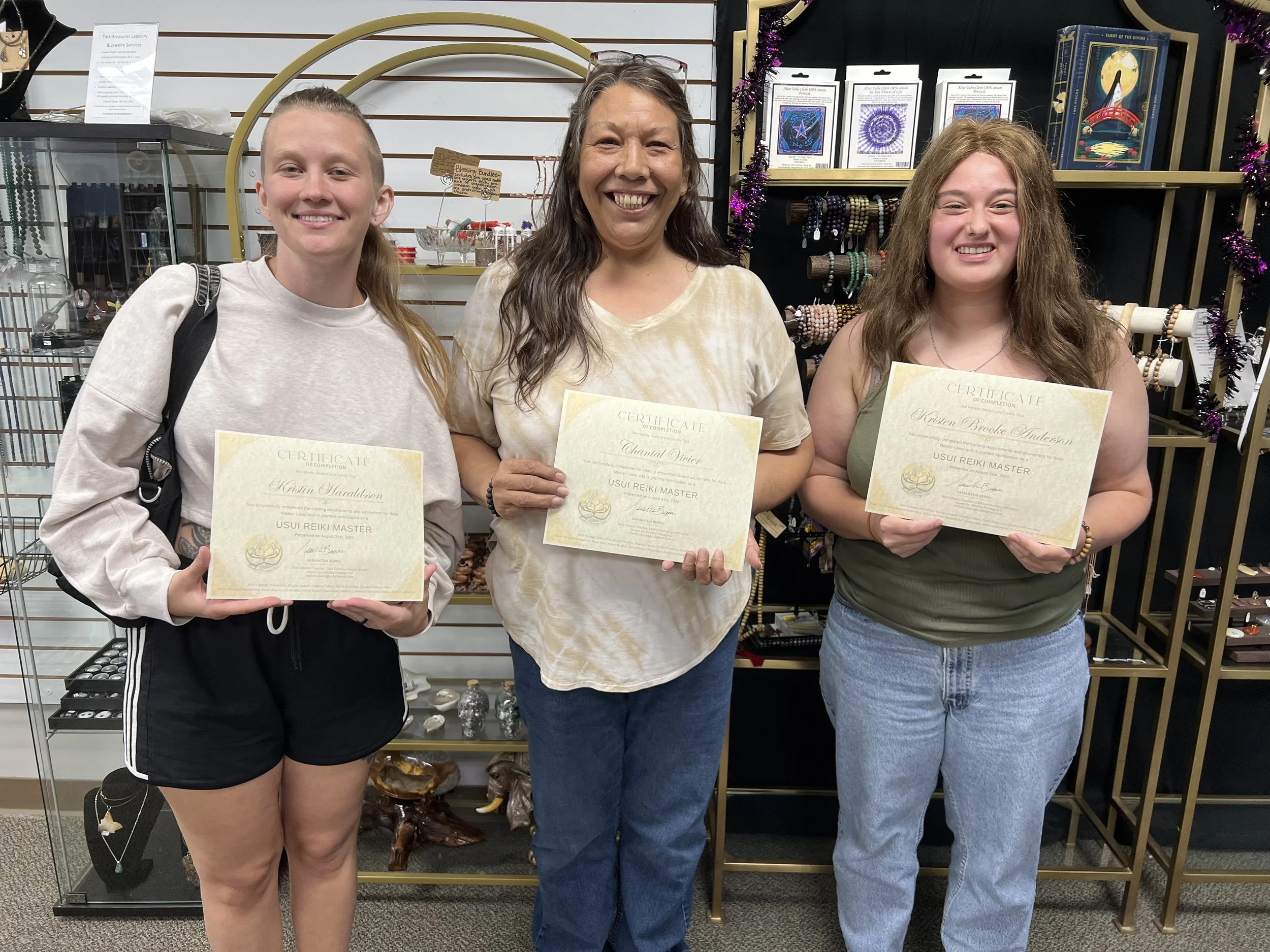 Three women standing indoors, each holding a certificate of completion for a Usui Reiki Master course, smiling at the camera. The woman on the left wears a light-colored sweatshirt and black shorts, the woman in the middle wears a tie-dye yellow and white shirt and jeans, and the woman on the right wears a green sleeveless top and jeans. Behind them are display shelves with jewelry and decorative items.