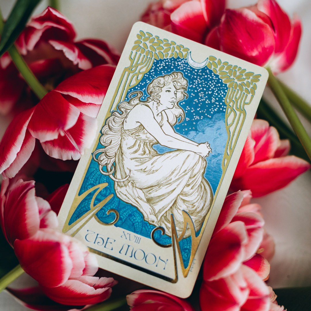 Tarot card titled 'The Moon' featuring an illustration of a woman with long flowing hair sitting among clouds, with stars and a crescent moon in the background, surrounded by red and white tulips.