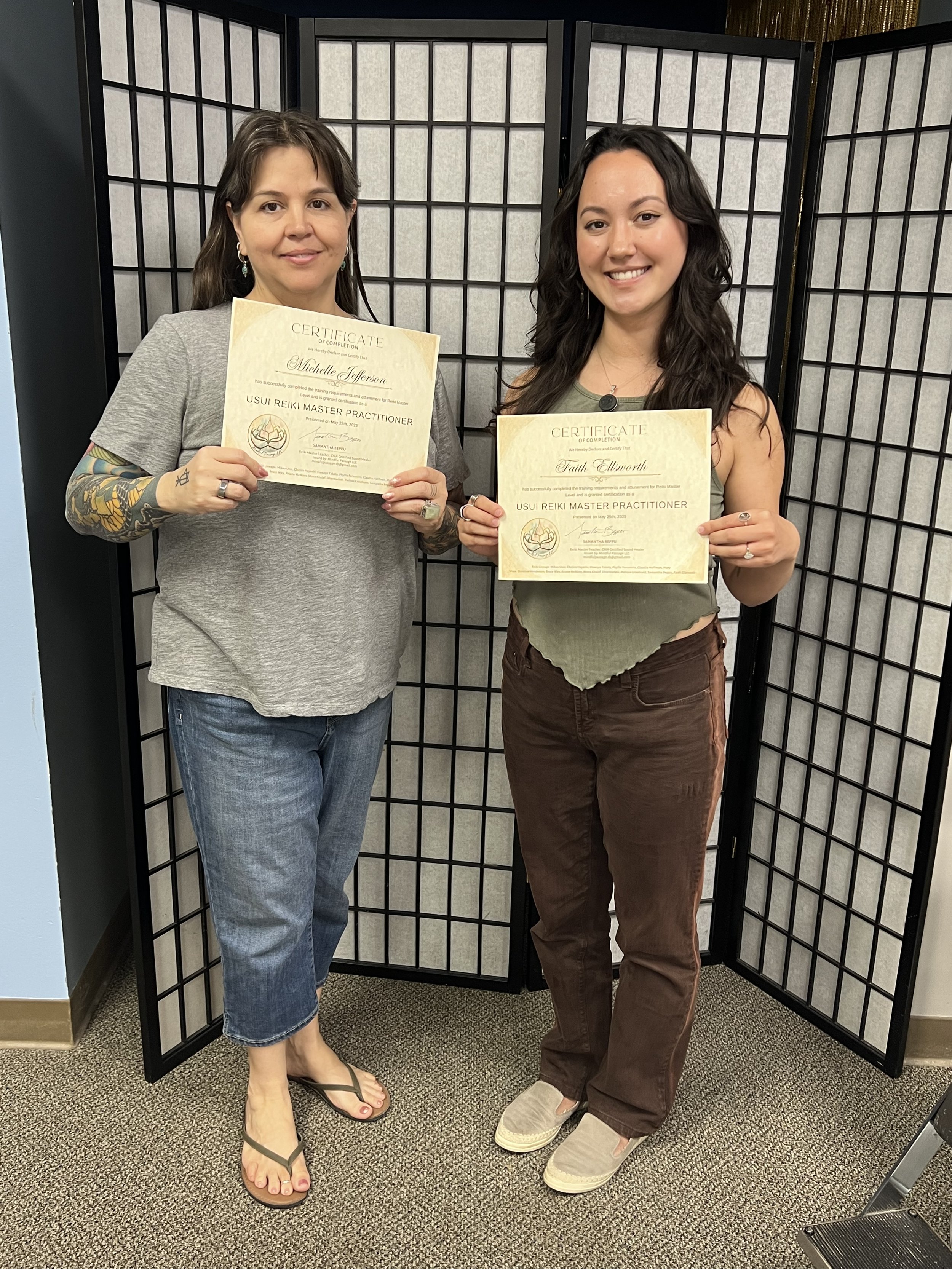 Two women standing in front of a room divider, holding certificates of completion for USUI Reiki Master Practitioner. The woman on the left has brown hair, tattoos on her right arm, and is wearing a gray t-shirt and blue jeans. The woman on the right has black hair, and is wearing a green sleeveless top and brown pants.