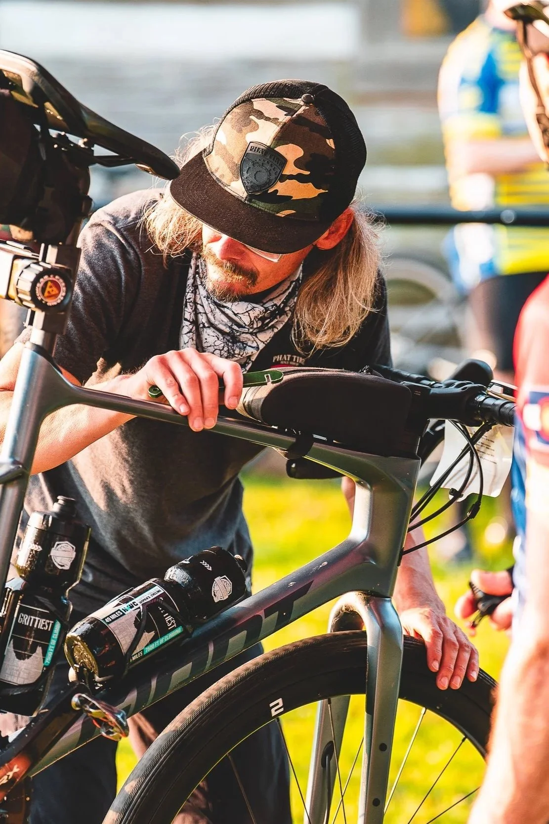 A man with long blonde hair, glasses, and a camouflage cap works on a mountain bike outdoors with a person nearby and parked cars in the background.