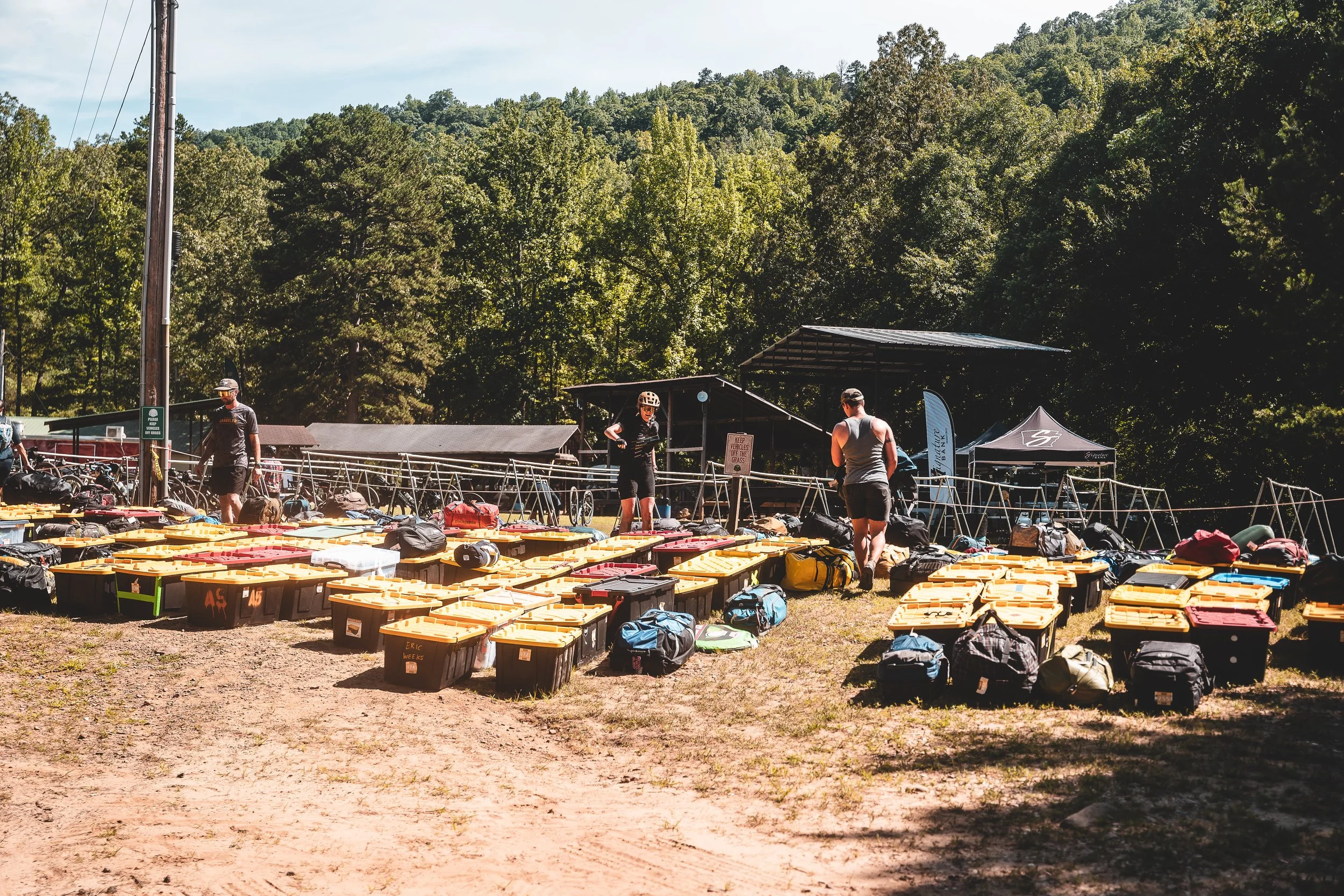 Outdoor scene at a cycling event with numerous open storage containers and gear laid out on the ground, several people preparing or organizing, tents in the background, and a lush green forested hillside in the distance.