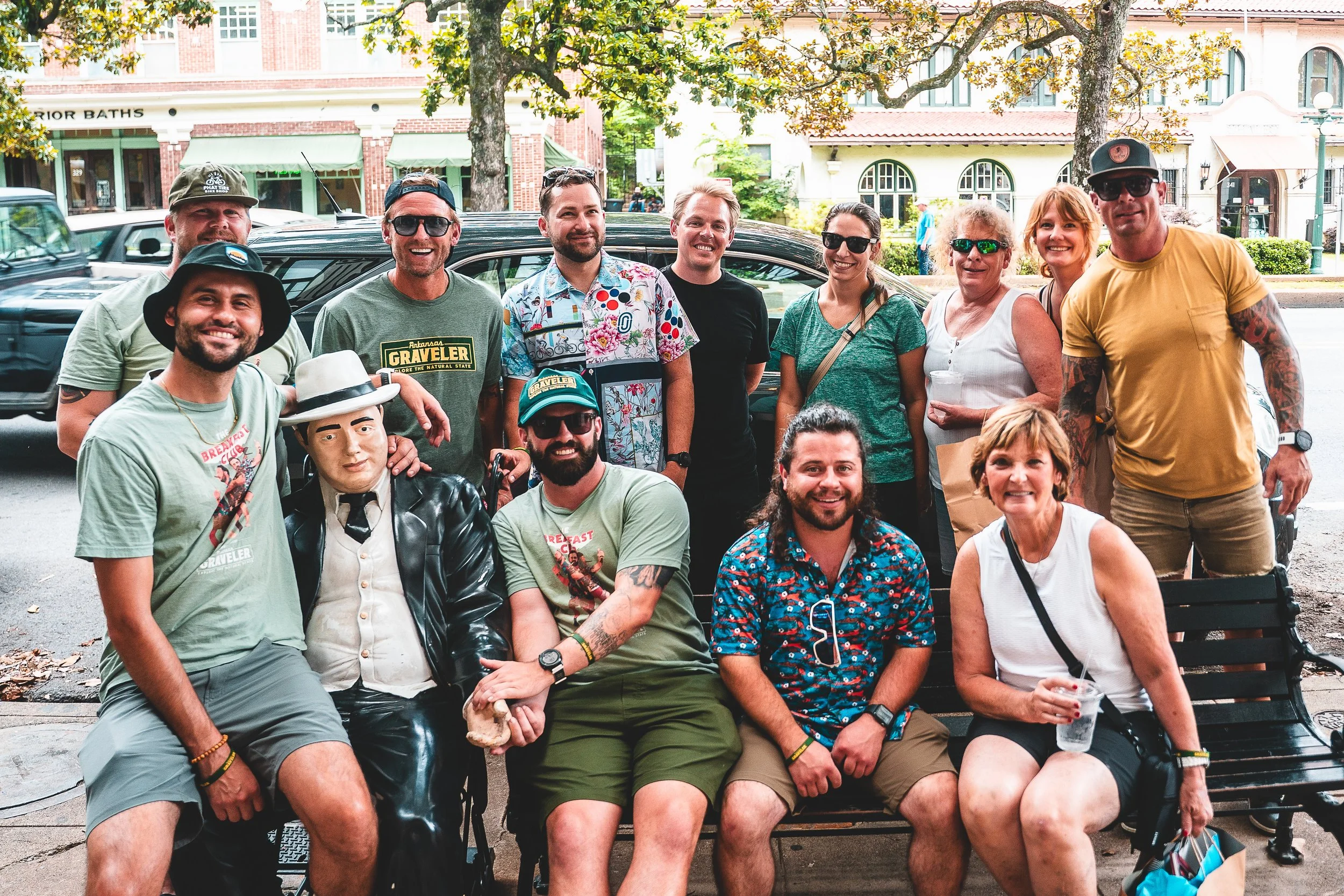 A group of people posing for a photo on a park bench, smiling and dressed casually under trees with buildings in the background.
