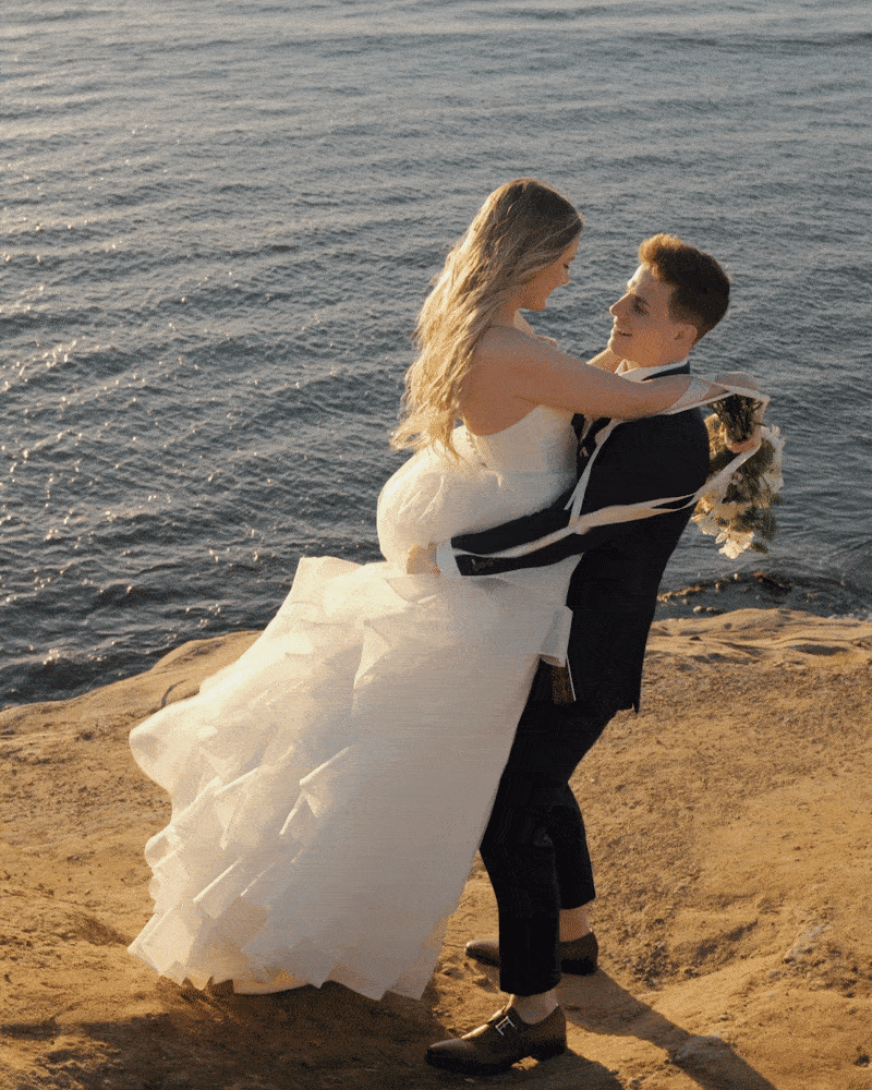 A couple dressed in wedding attire, with the groom in a black suit and the bride in a white wedding dress, embracing by the water on a rocky shoreline during sunset.
