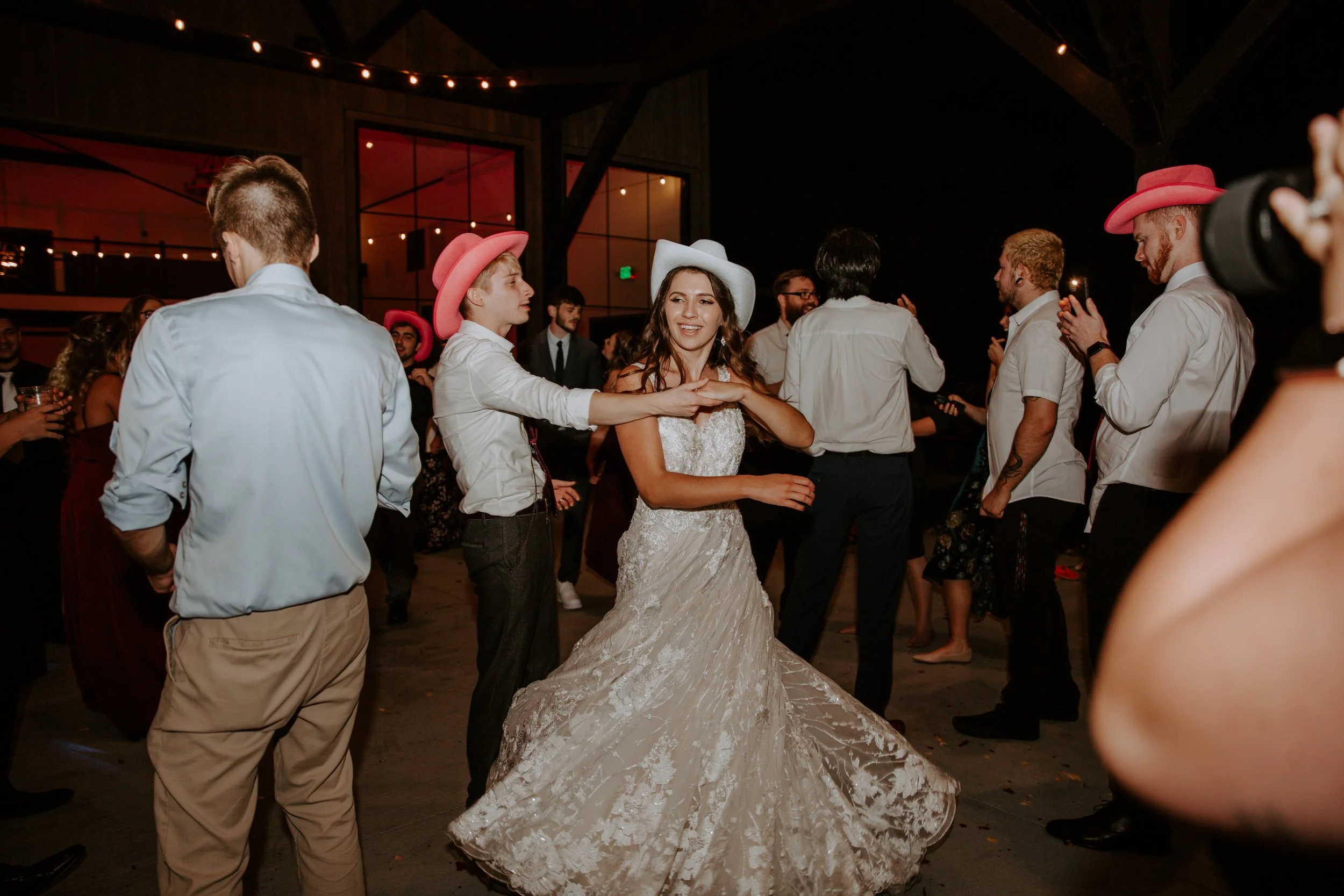 Bride and groom dance together under string lights outdside of their wedding reception, both wearing cowboy hats.