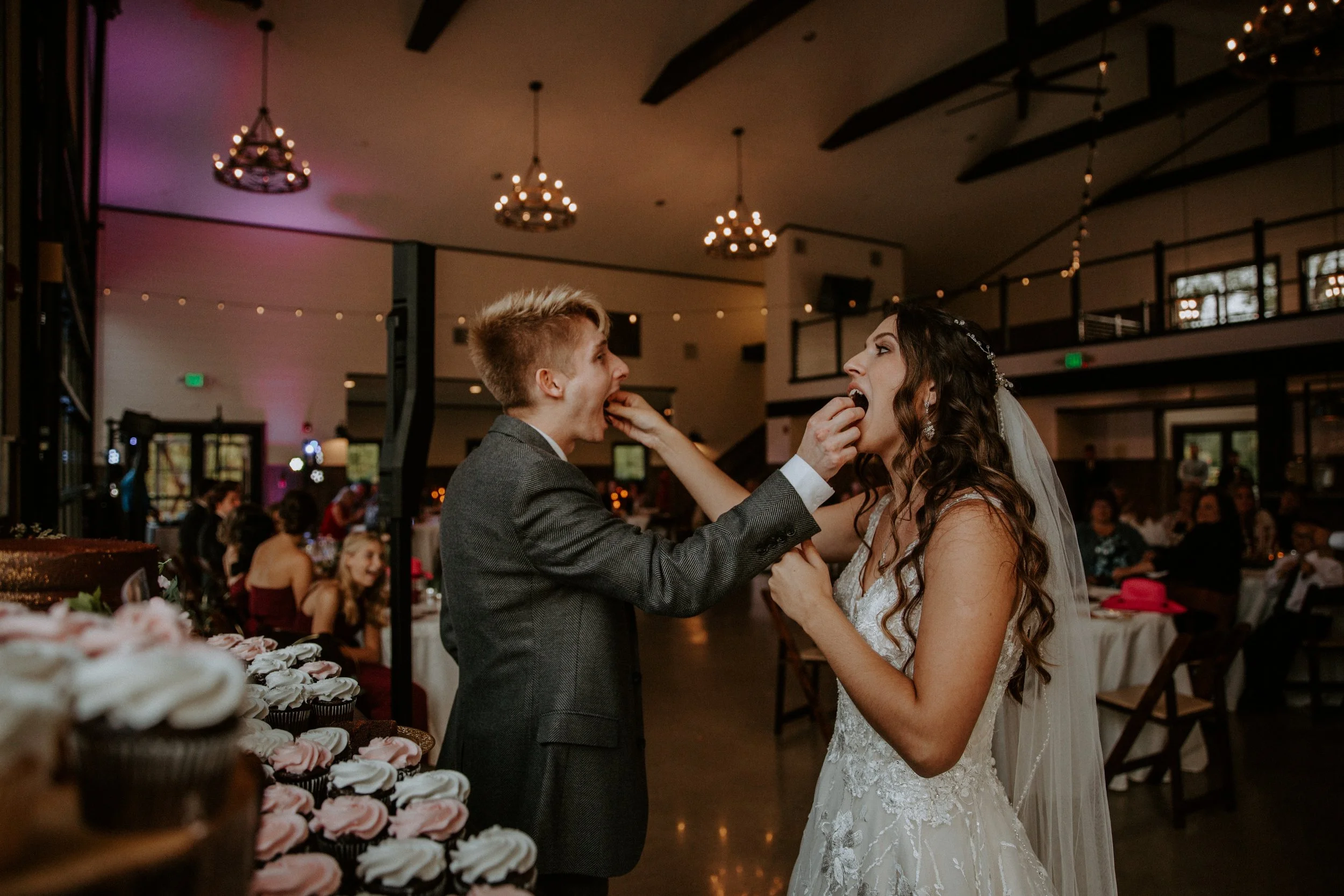 Bride and groom feed each other cupcakes during their wedding reception, surrounded by a table of desserts.