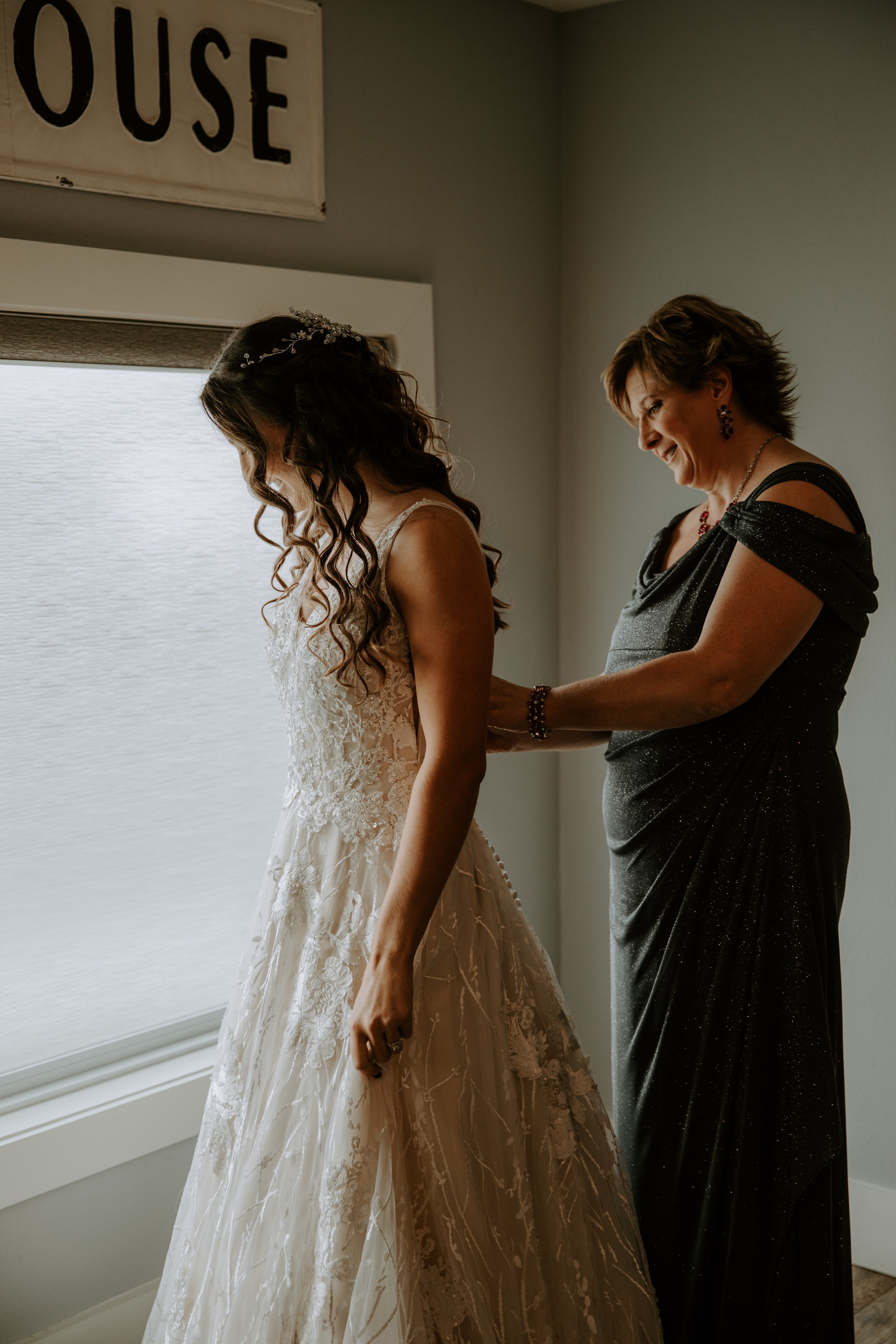 Bride getting ready with help from a loved one, smiling softly as she looks out the window.