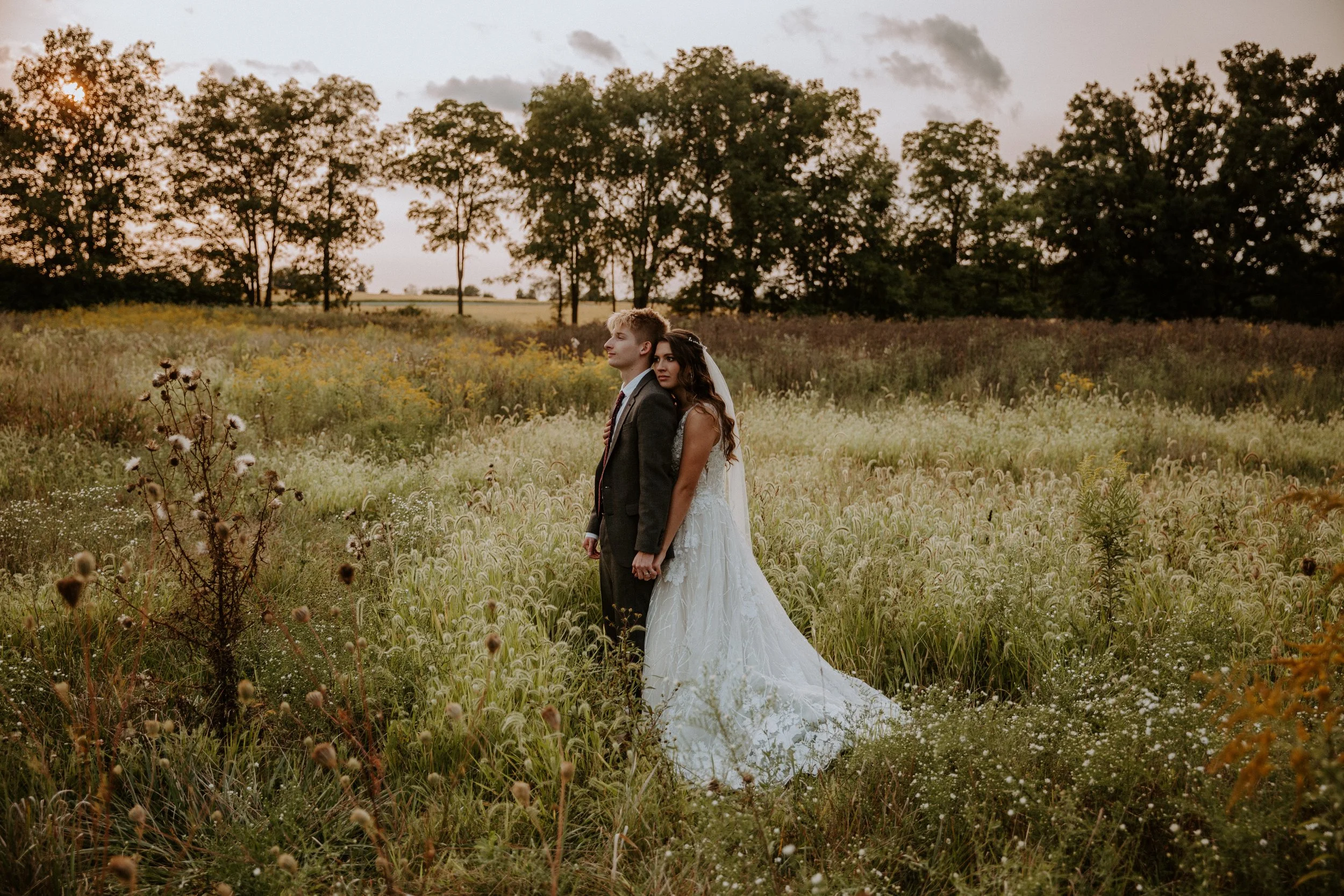 Bride and groom stand together in a field, holding hands and looking in opposite directions as the sun sets behind them.