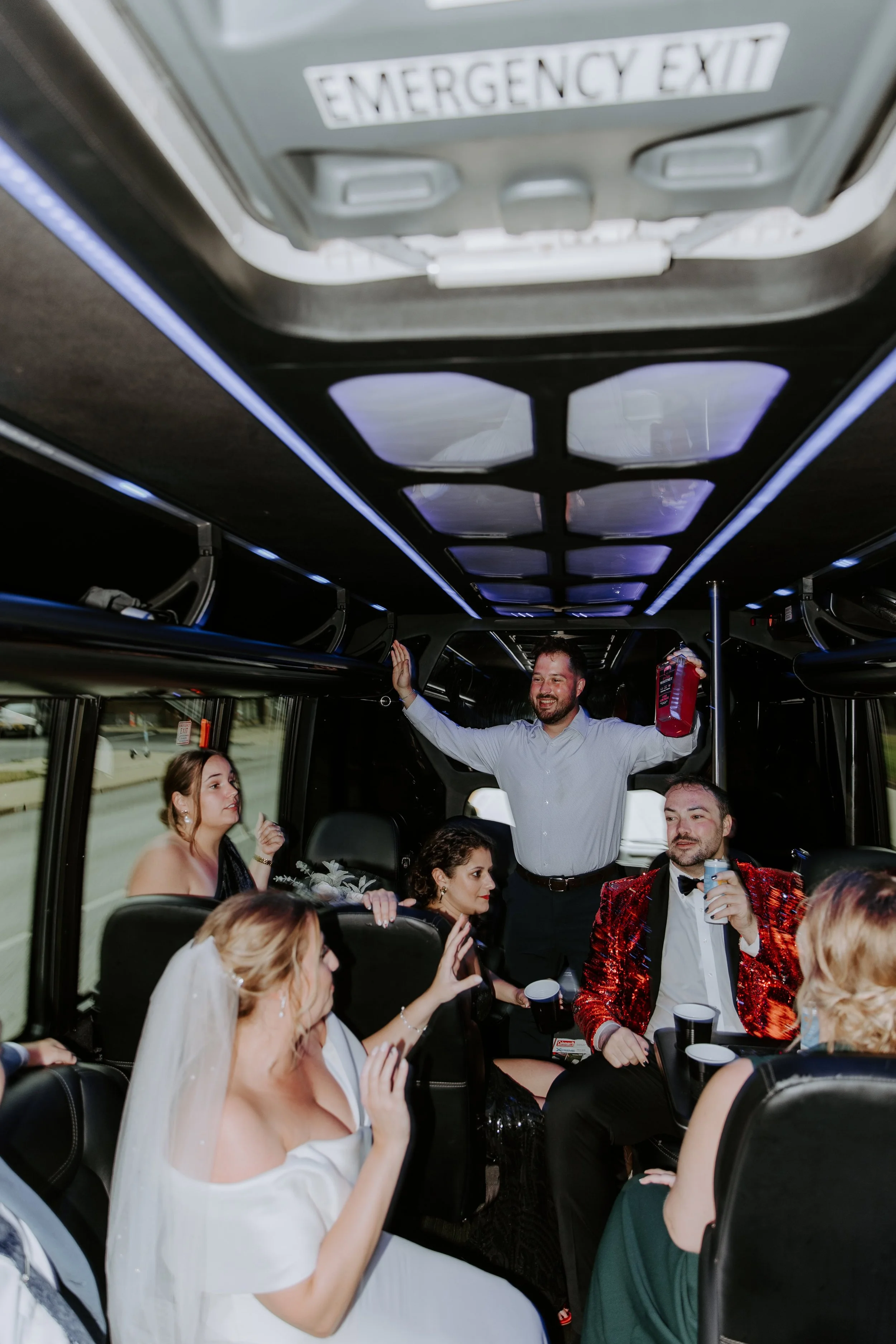 A man wearing a blue button up shit and black pants holds up a large bottle of Pink Whitney while standing in the middle of a party bus surrounded by friends.