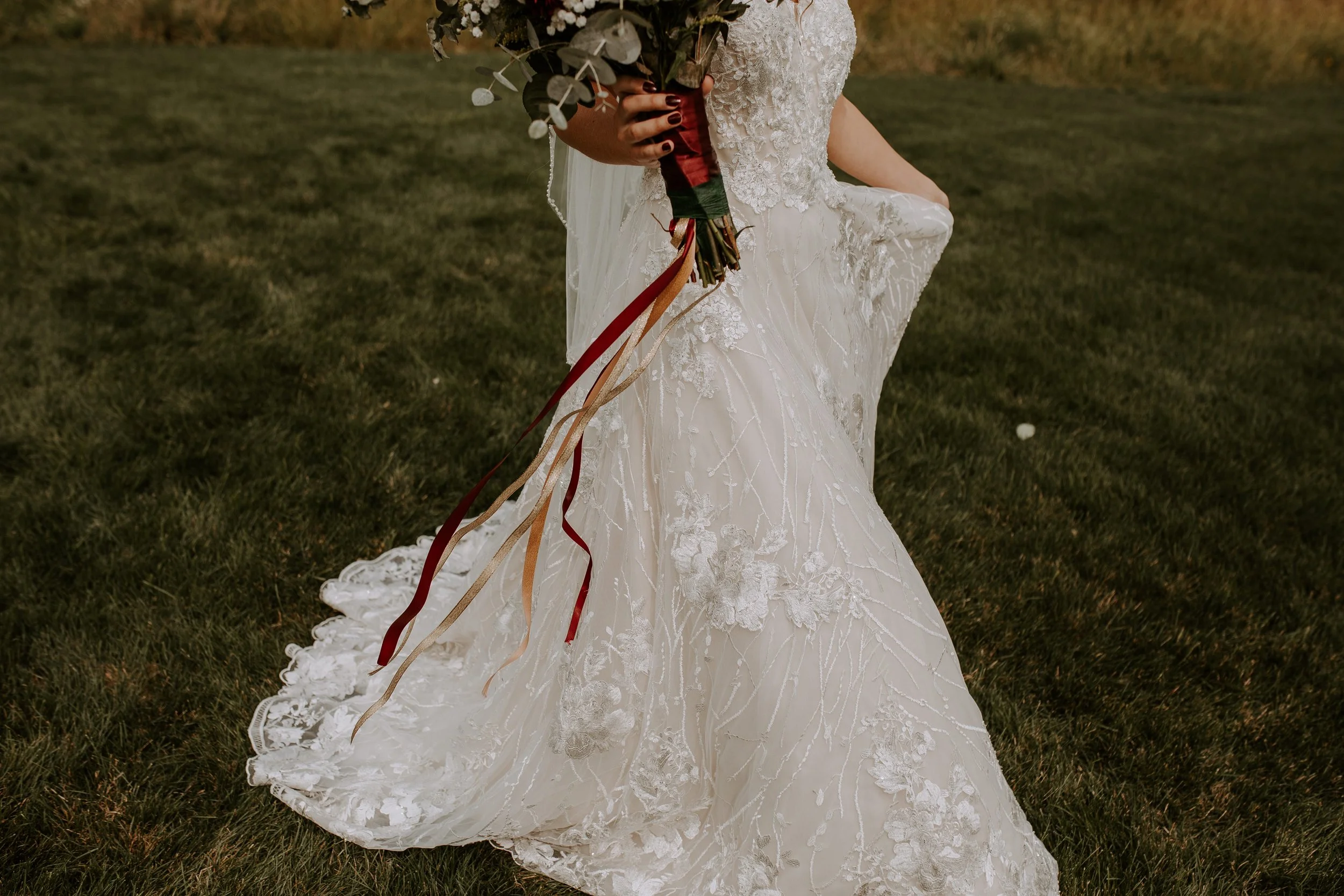 Bride lifting her wedding dress slightly while walking through a grassy field, showing off the lace details and ribbons trailing from her bouquet.