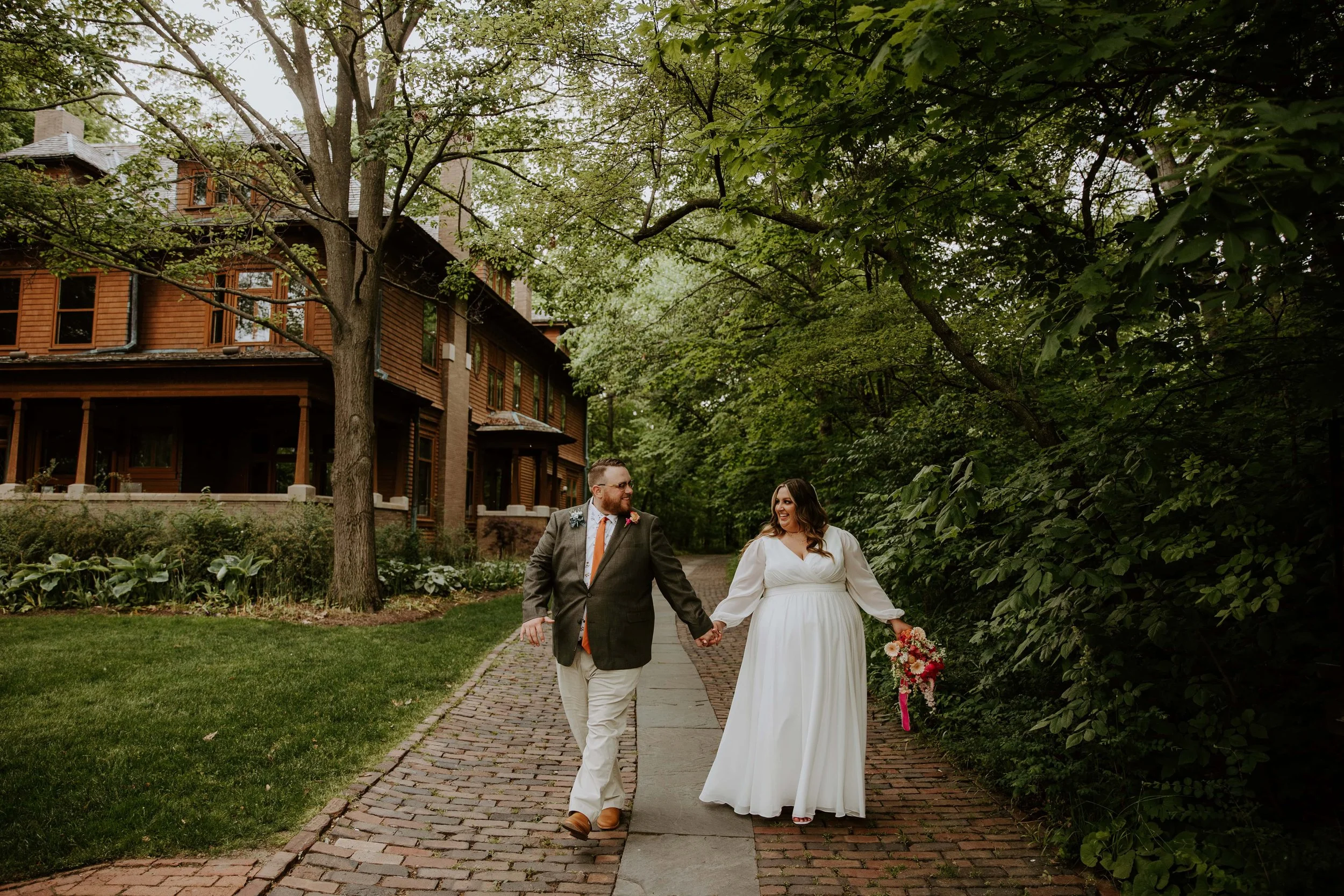 The groom and bride hold hands and walk along the brick path next to the Oakhurst House at Minnetrista.