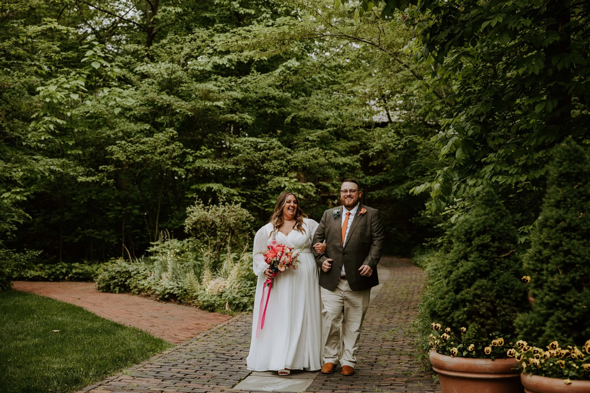 The couple walk arm in arm along a wooded brick path.