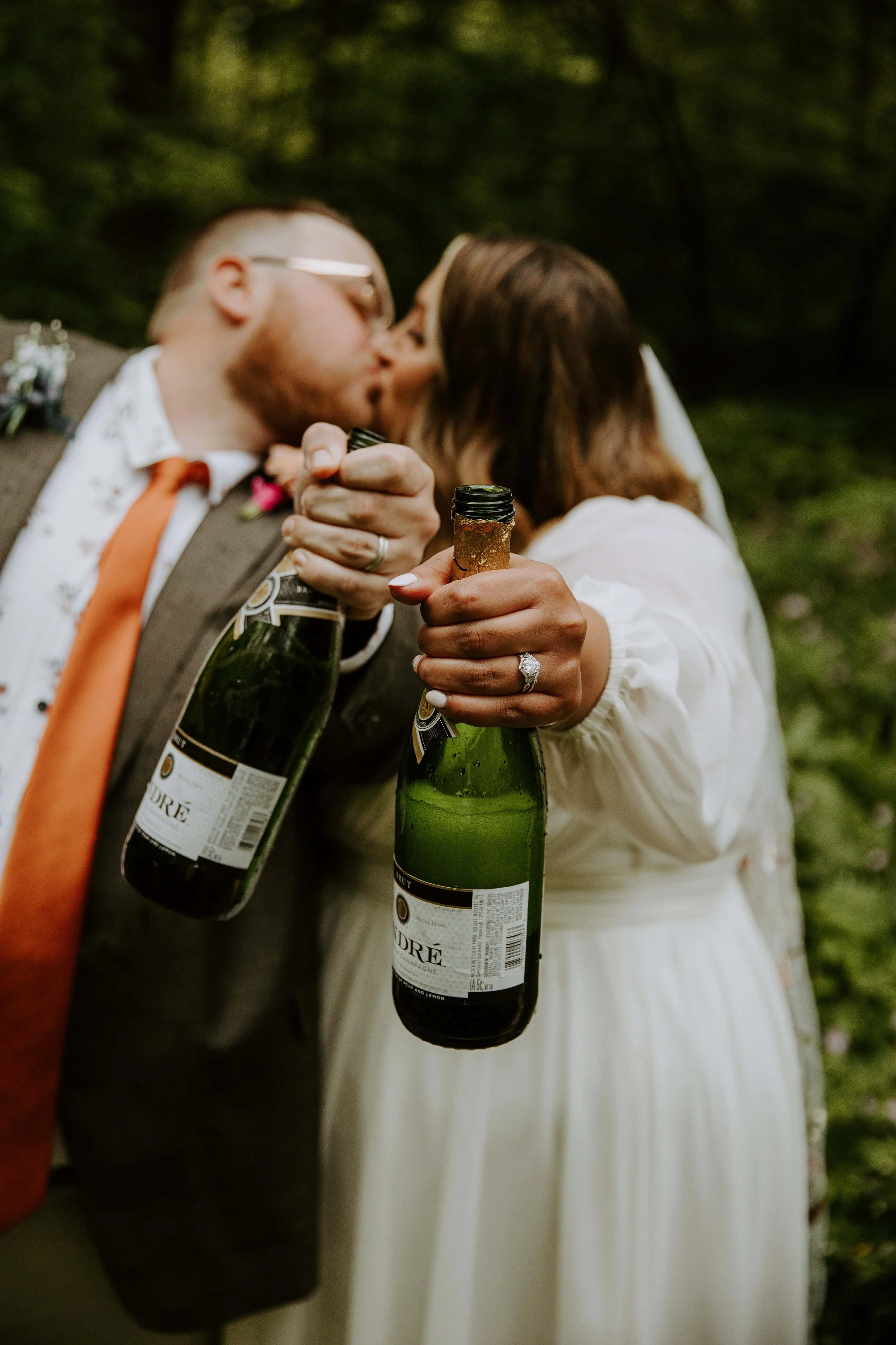 The bride and groom kiss in the background with their arms extended to show off their wedding rings and champagne bottles.