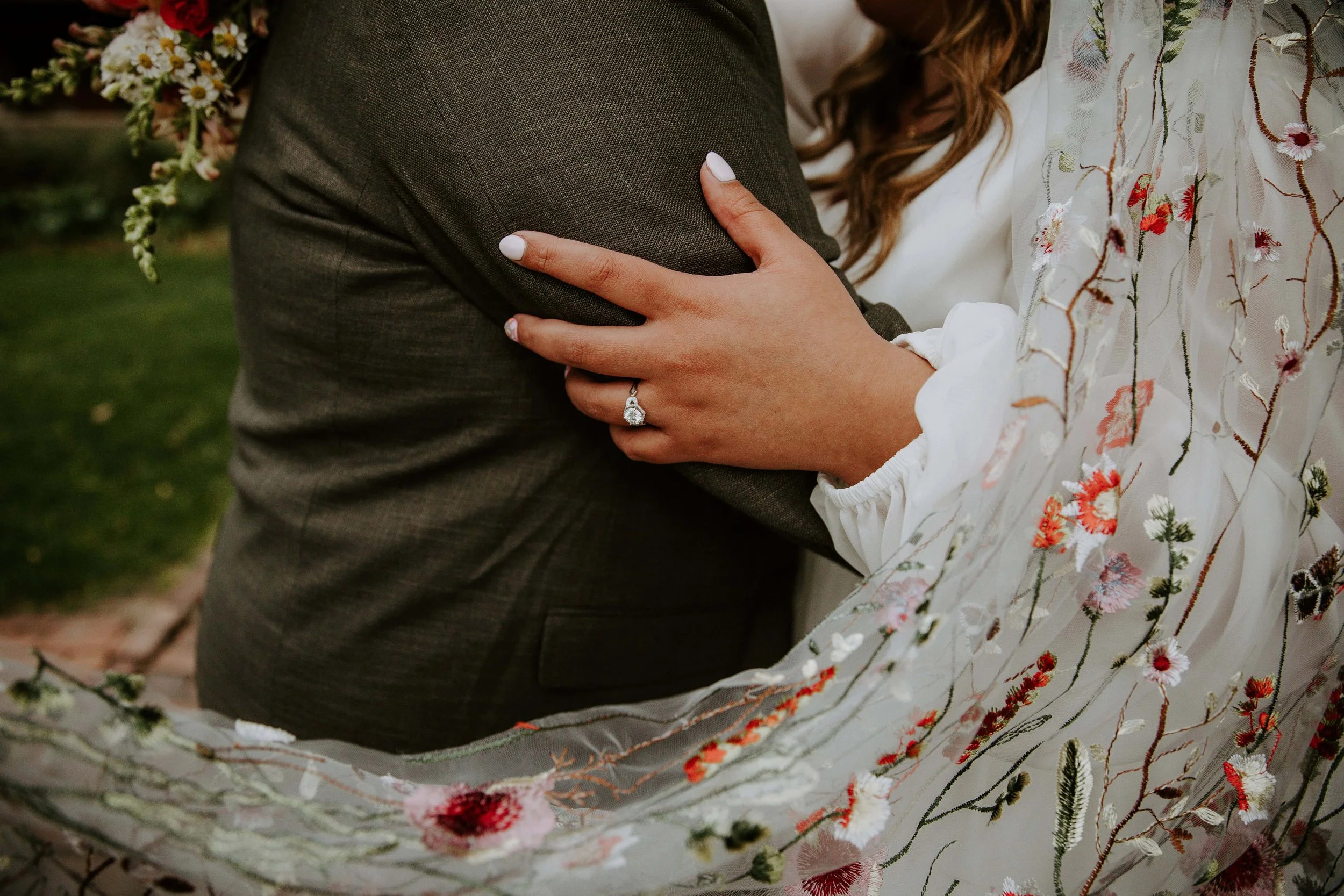 A close up shot of the bride's engagement ring while she rests her hand on the grooms arm.
