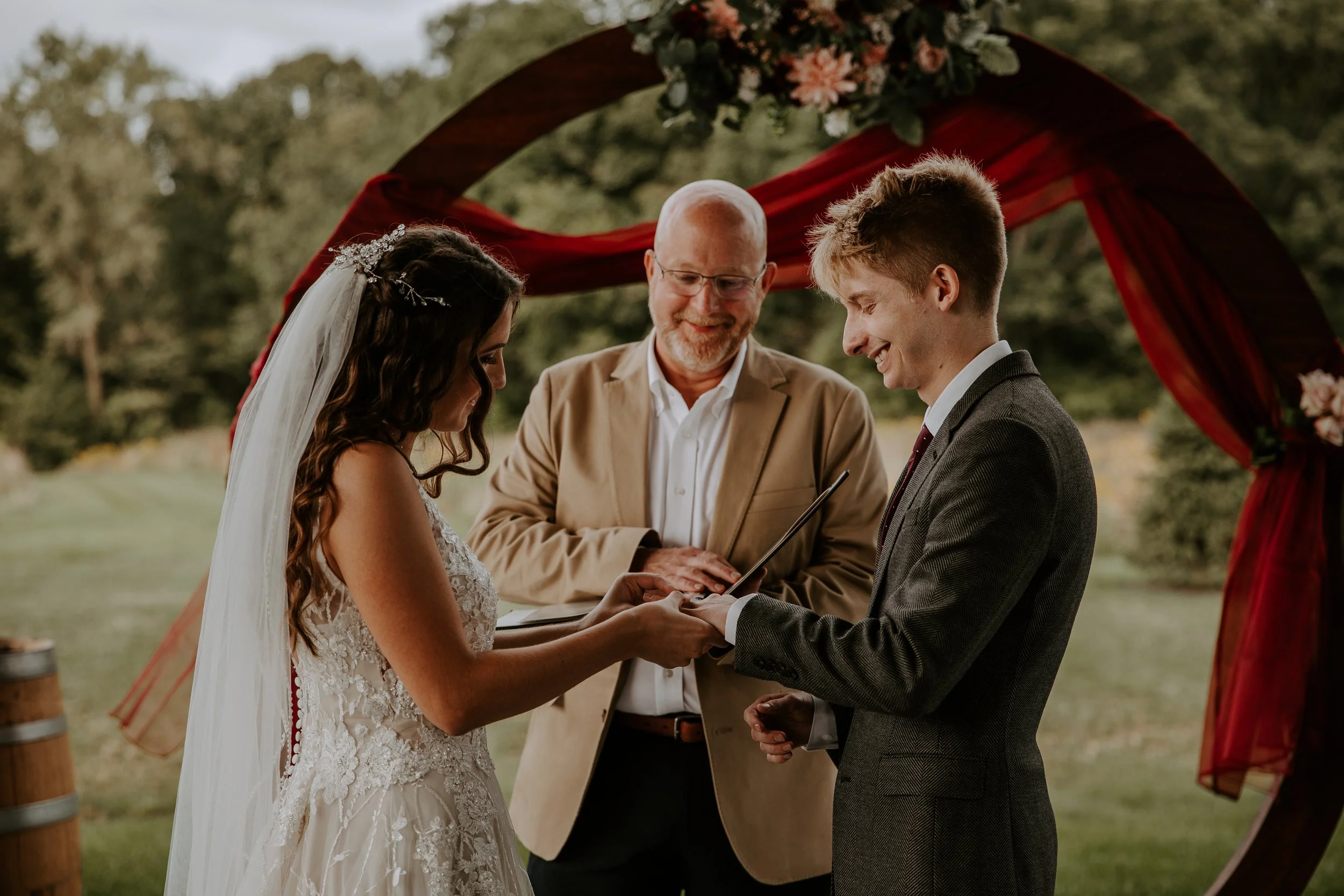 Bride and groom exchanging rings during their outdoor wedding ceremony, with the officiant smiling behind them under a decorated arch.