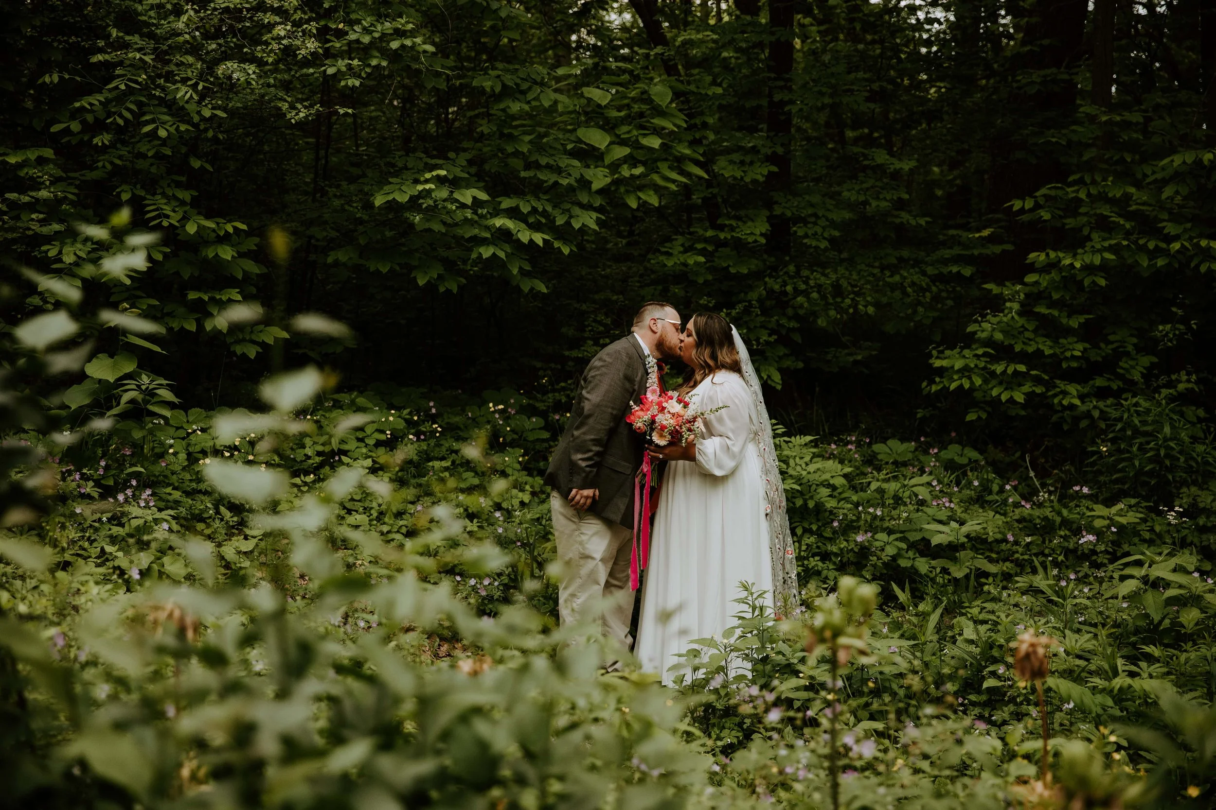 The couple kiss, surrounded by trees and greenery.