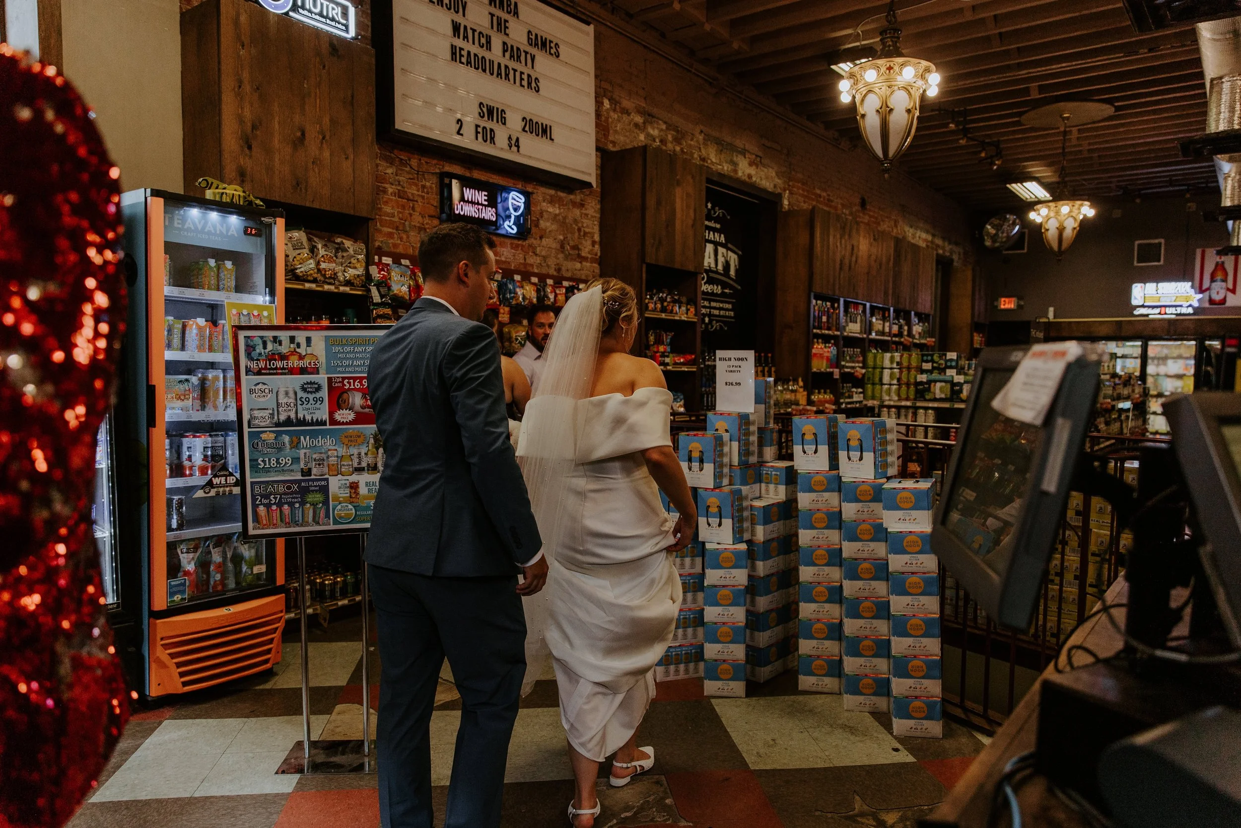 A bride and groom in their wedding attire walk around a liquor store.
