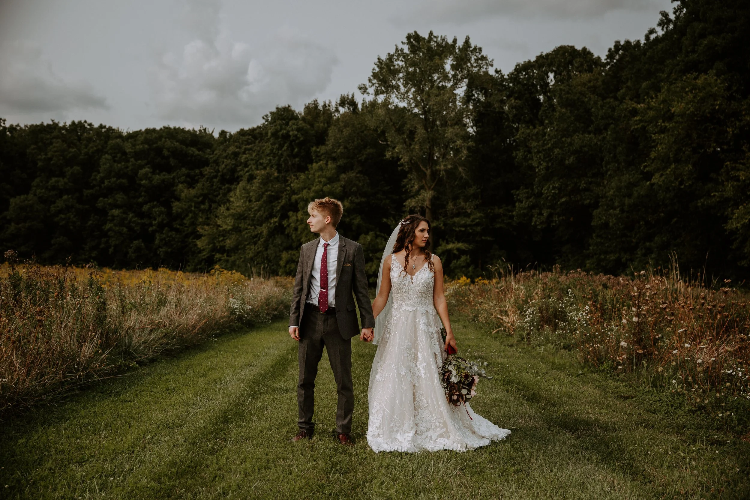Bride and groom hold hands while standing in a grassy field, gazing off into the distance.