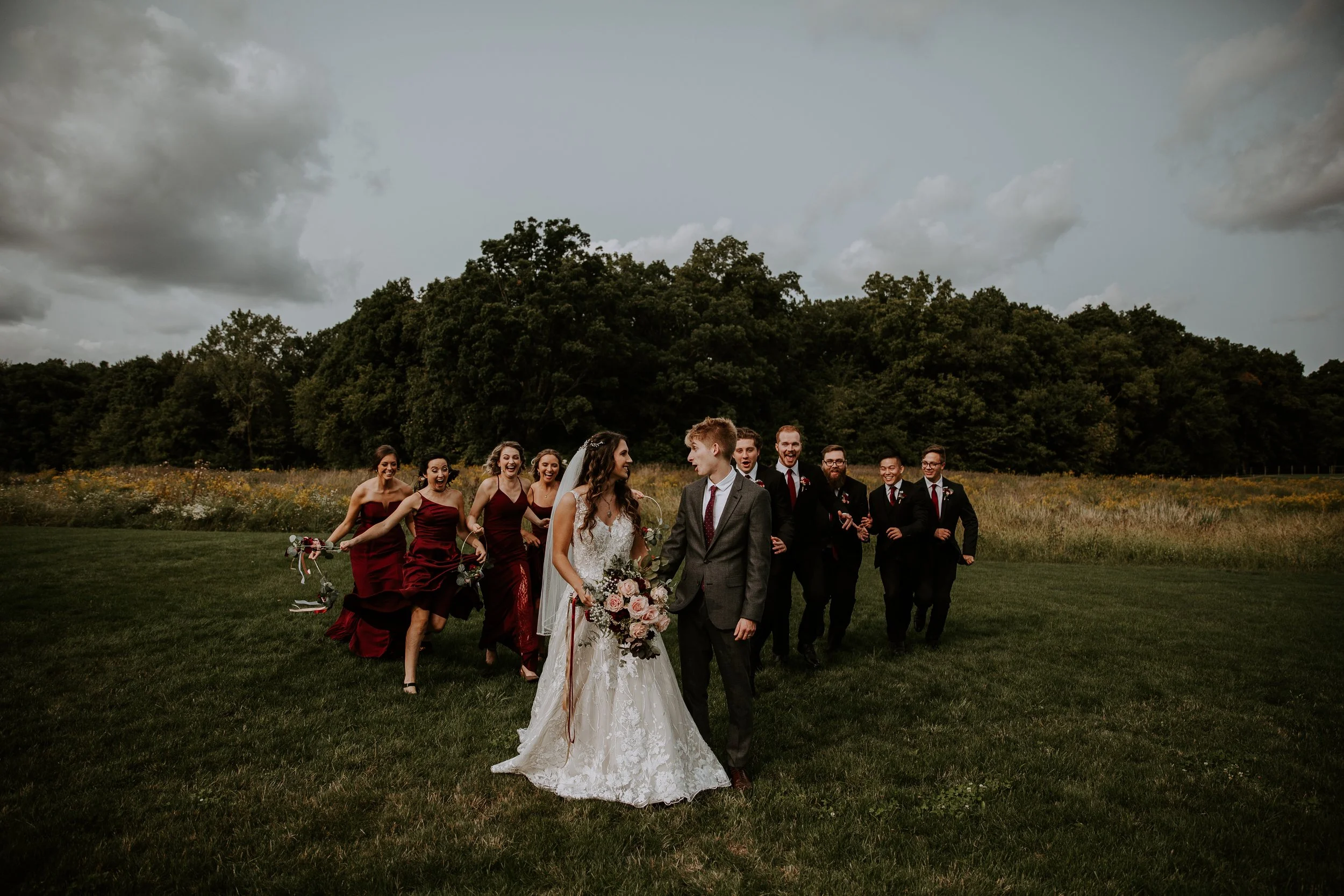 Bride and groom walking hand in hand, smiling as their wedding party follows behind them through an open field.