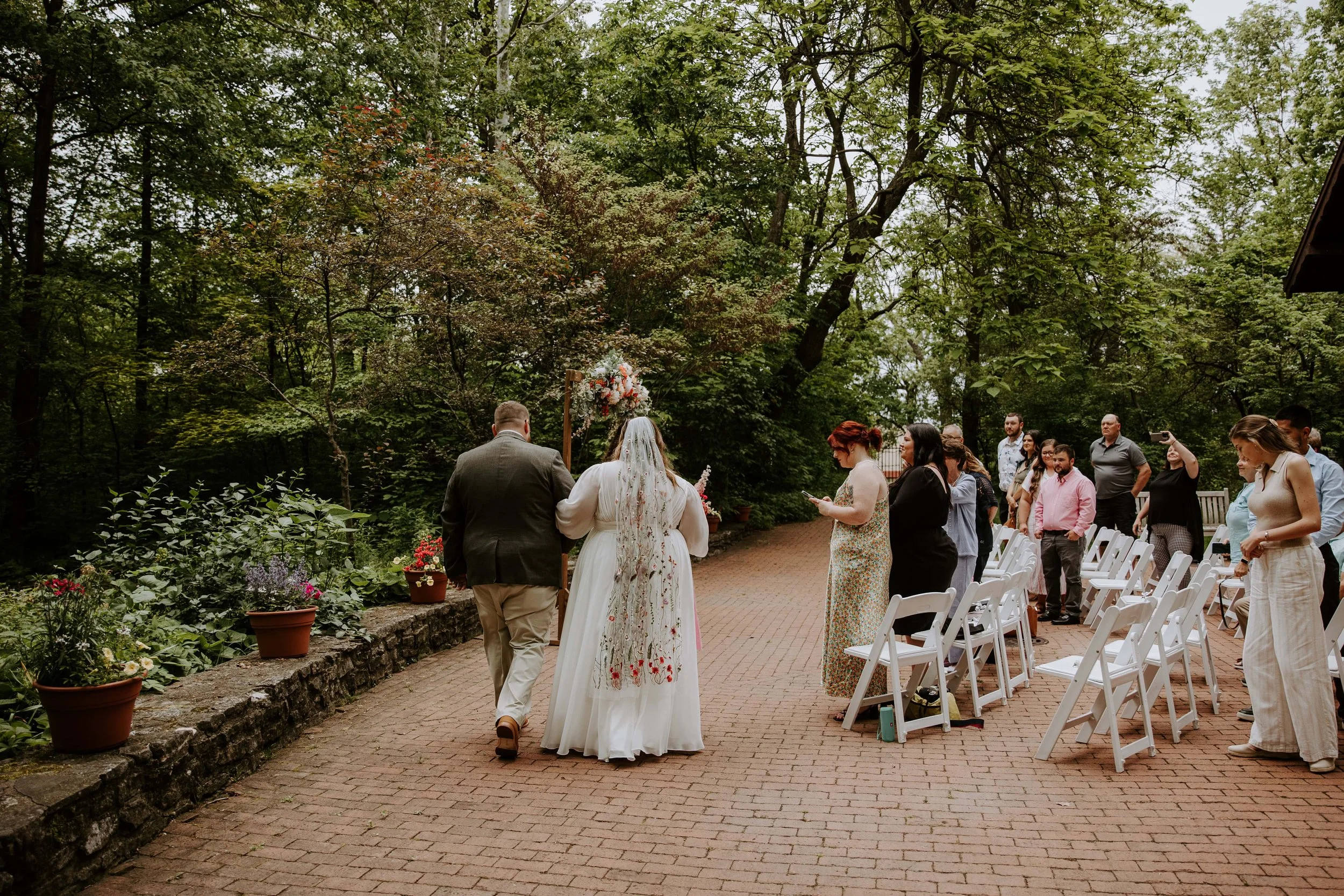 The bride and groom with their backs to the camera walk to their ceremony arch. Their guests are standing at their seats to the right.