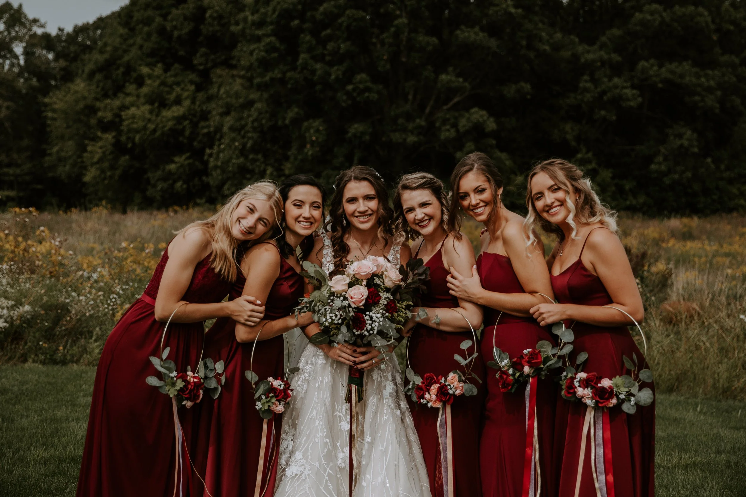 Bride and bridesmaids pose together, holding bouquets and smiling against a backdrop of greenery.