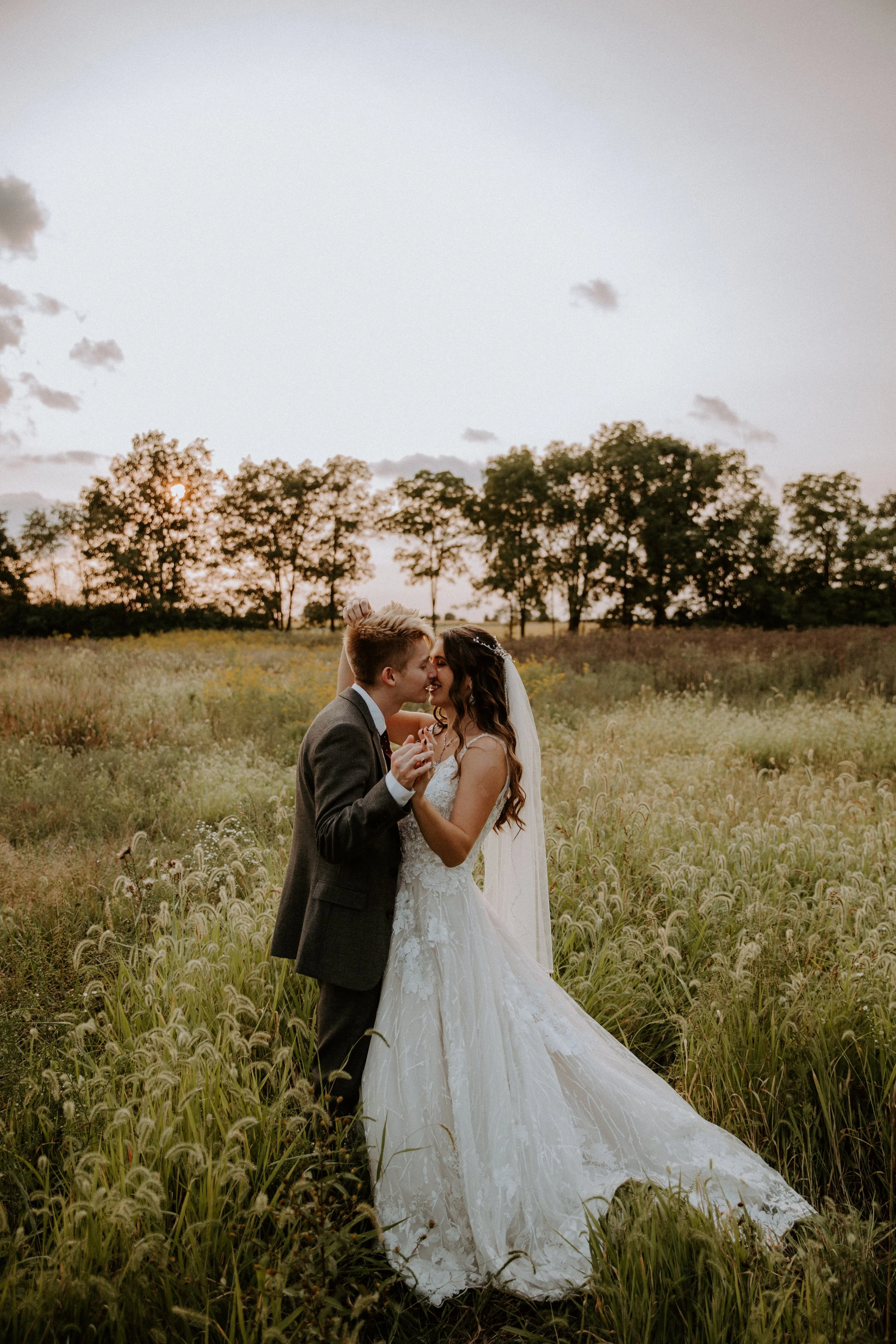Bride and groom kiss in a field as the sun sets, surrounded by tall grass and wildflowers.