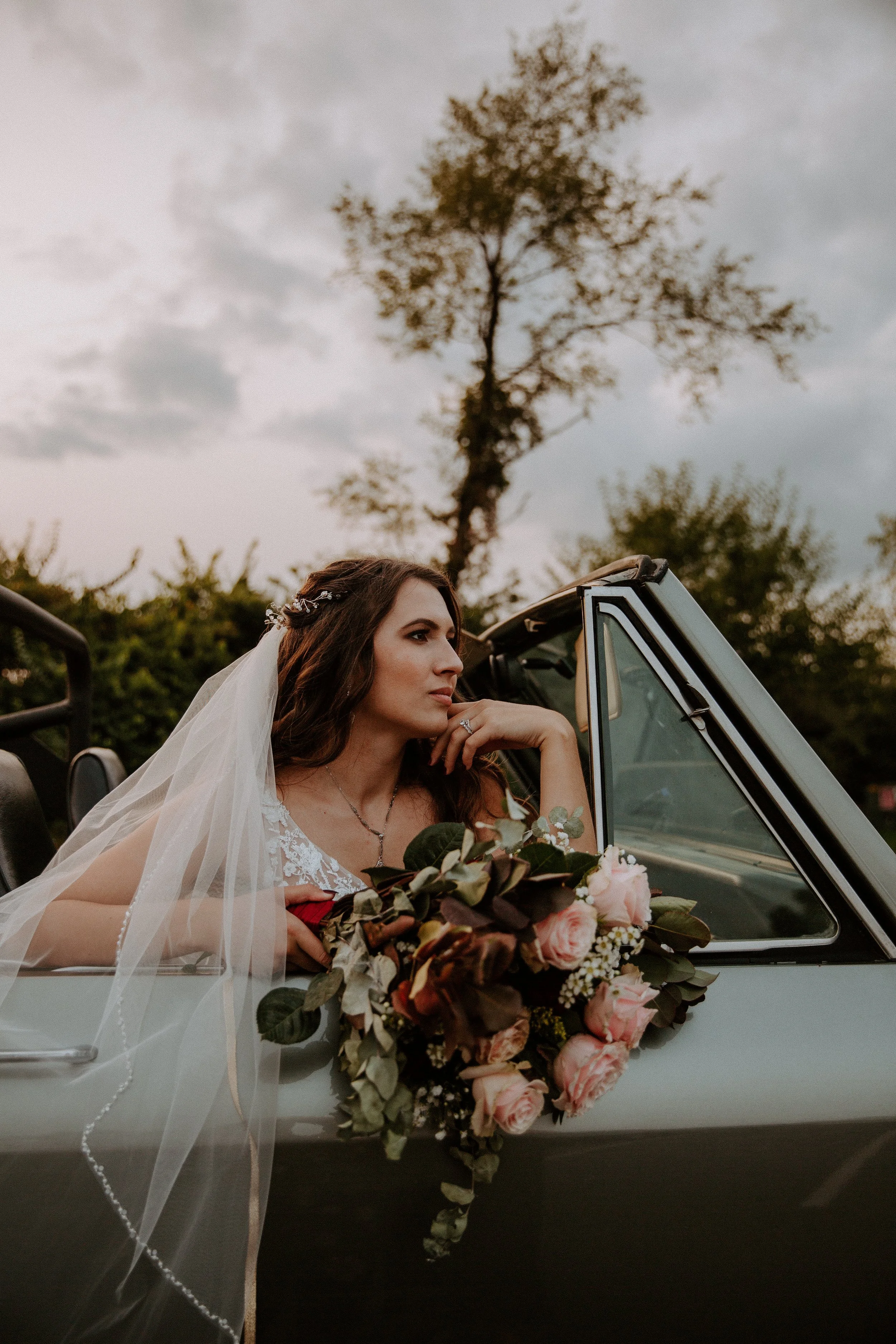 Bride sitting in a vintage convertible, holding her bouquet and gazing thoughtfully into the distance as the sun sets.
