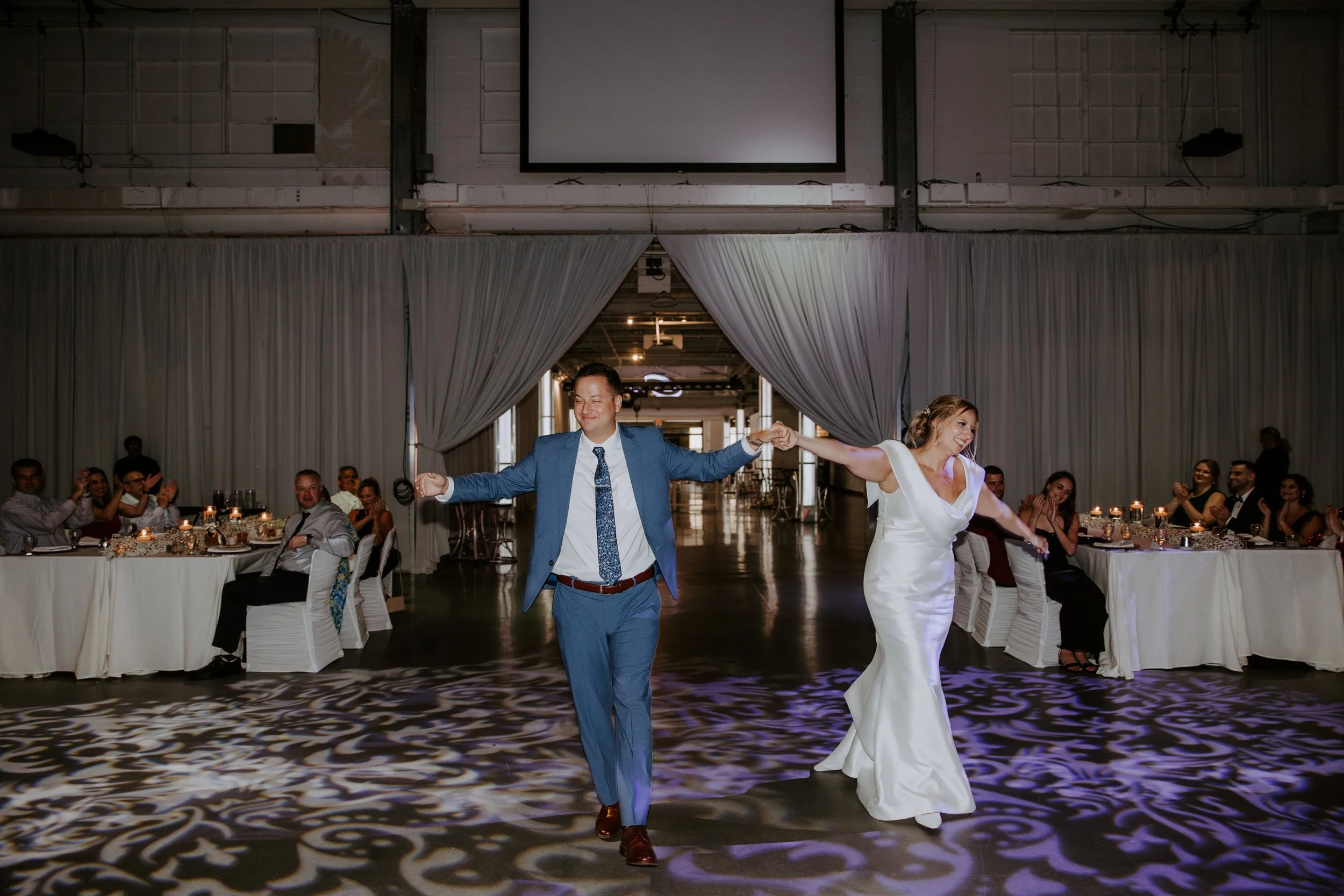 A bride and groom walk into their reception hand in hand with guests sitting at tables around them clapping their hands.