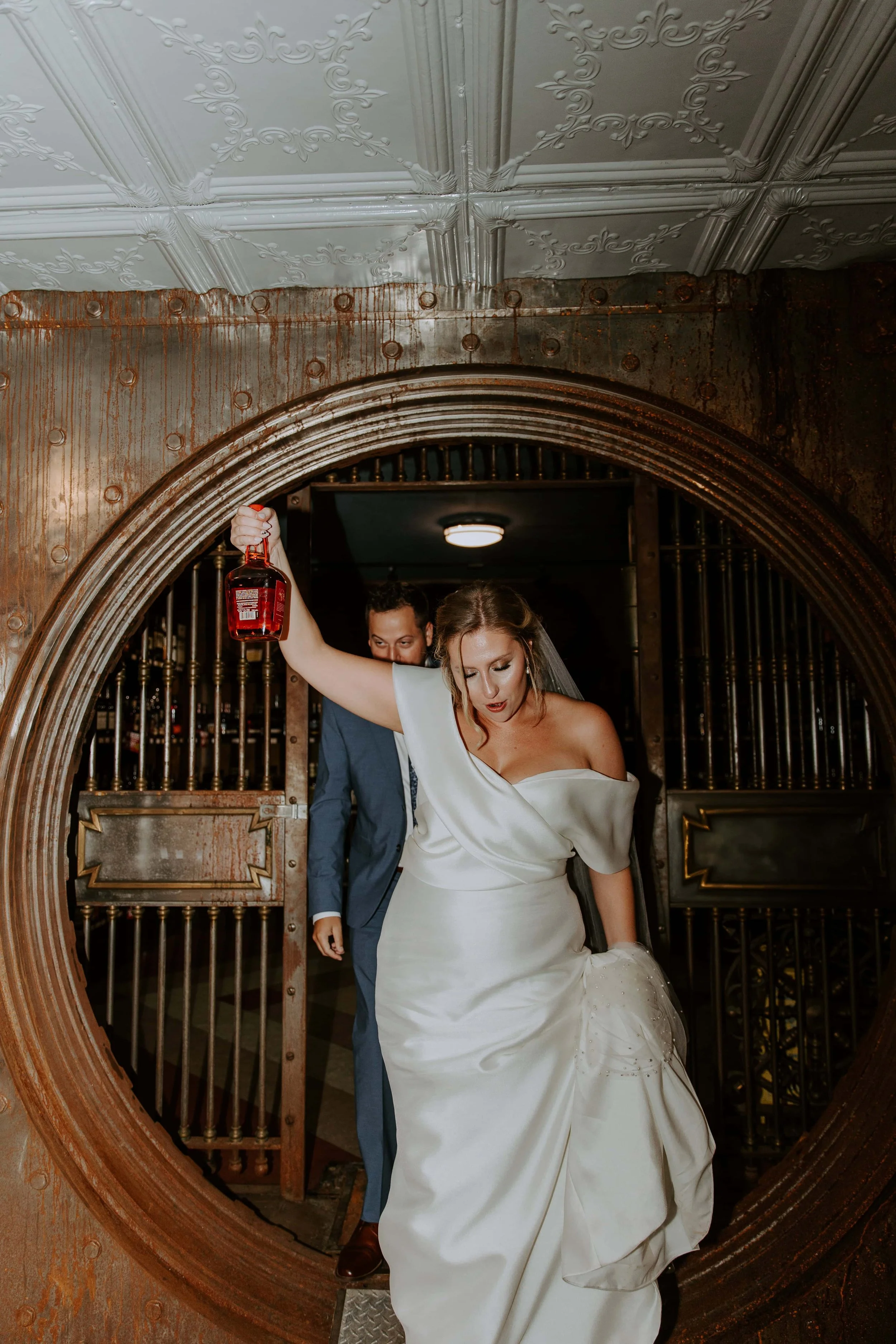 The bride and groom walk out of the whiskey vault. The bride is holding a bottle of whiskey up in the air.
