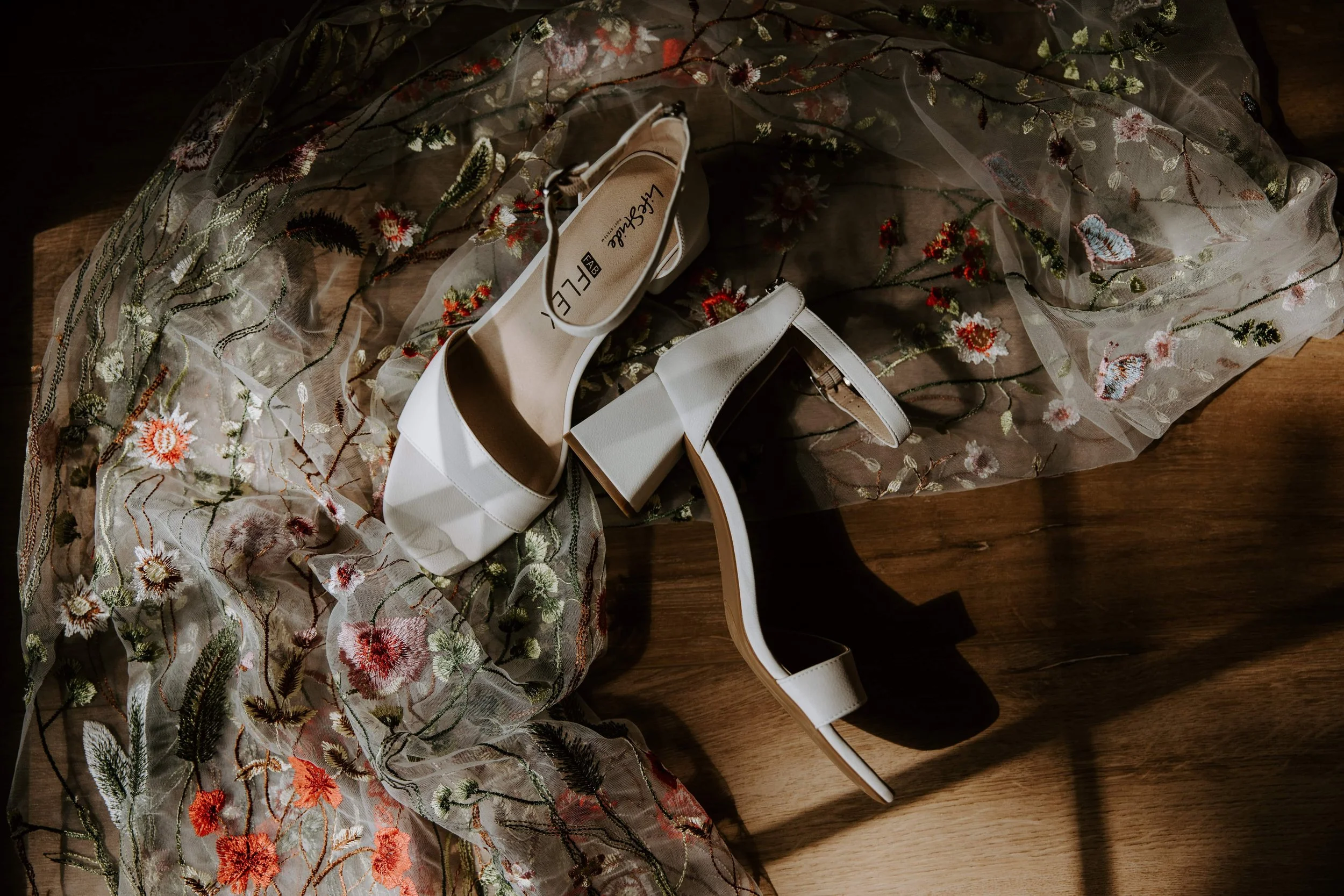 The sun shines through the window on a pair of white heels laying on a floral veil on the ground.