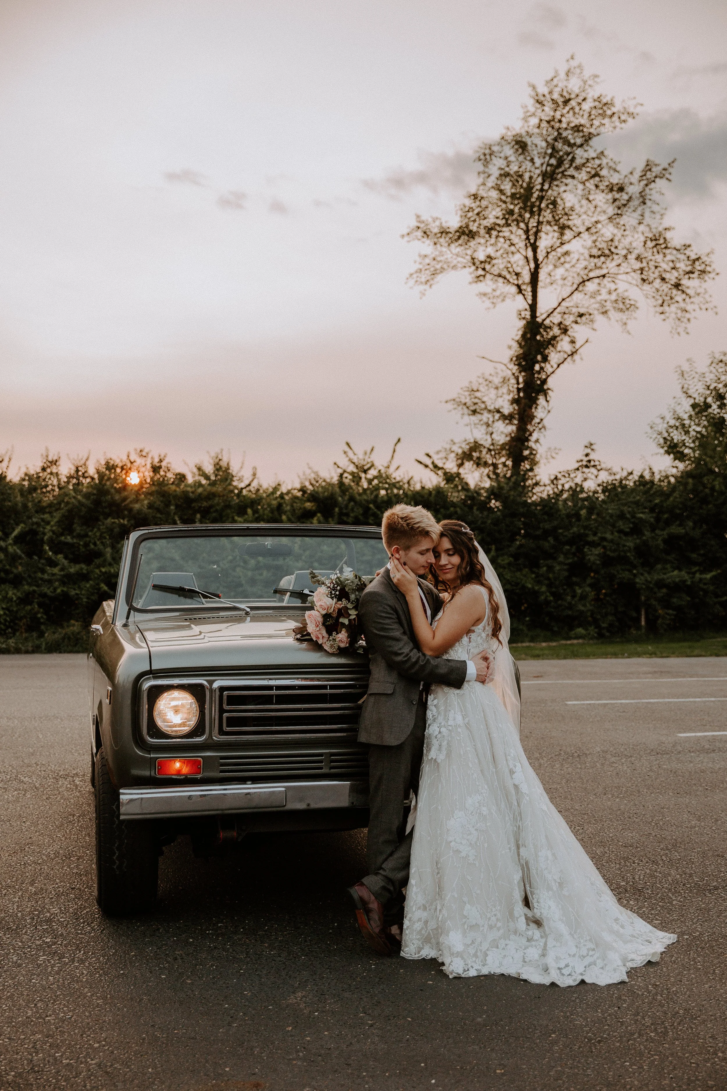 Bride and groom embrace in front of a vintage convertible at sunset, with the bride’s bouquet resting on the car’s hood.