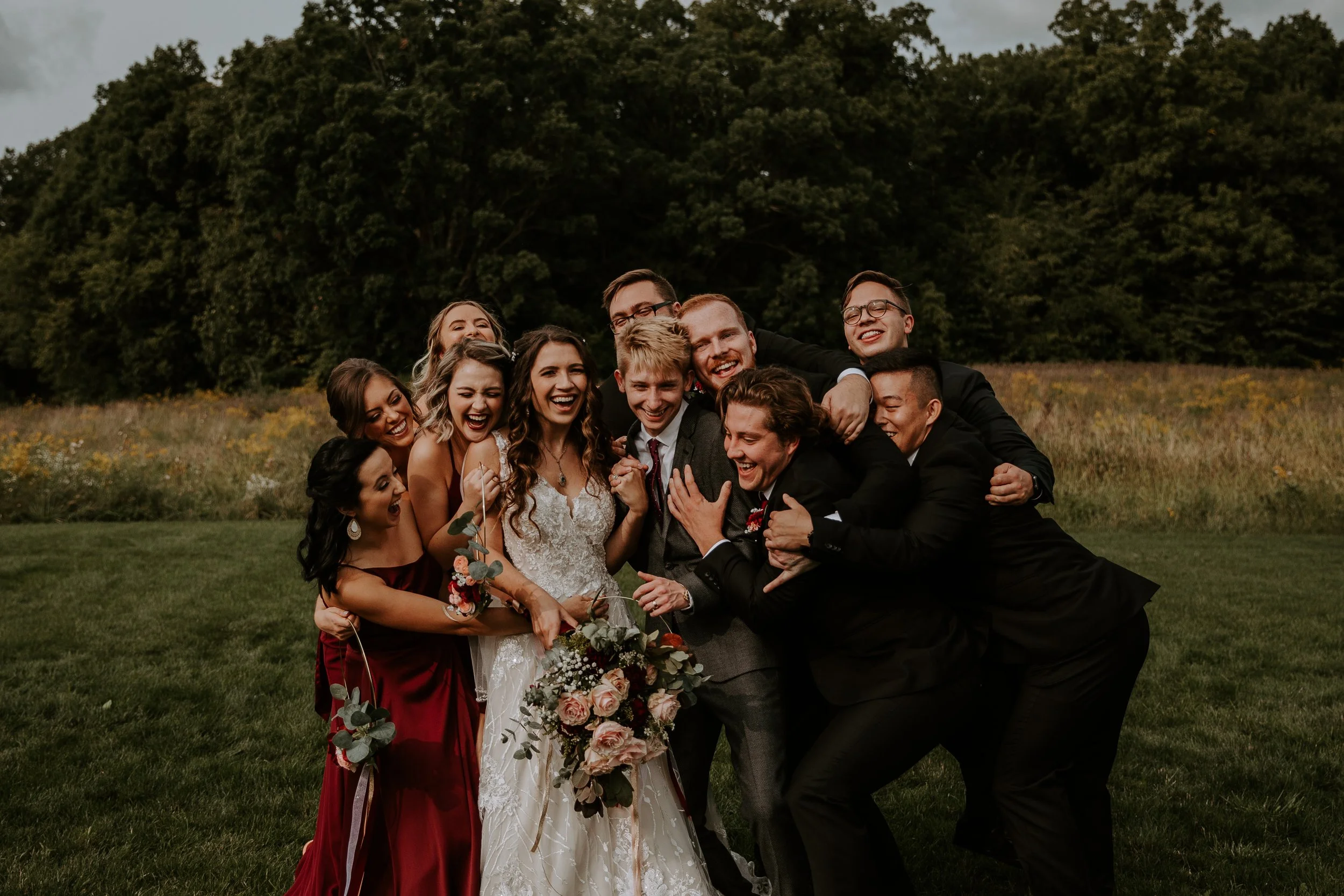 Bride and groom surrounded by their wedding party, all laughing and hugging in a joyful group embrace outdoors.