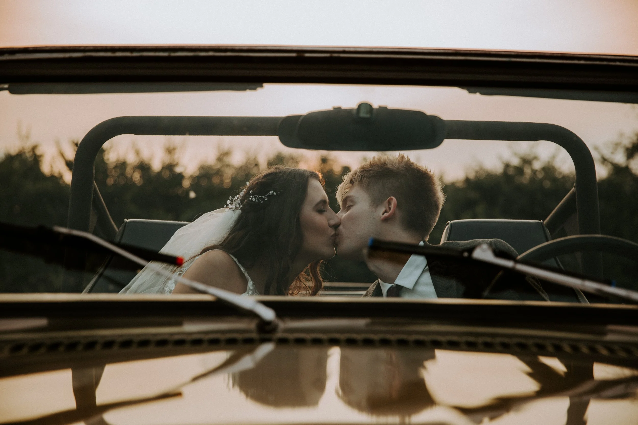 Bride and groom share a kiss inside a vintage convertible, viewed through the windshield at sunset.